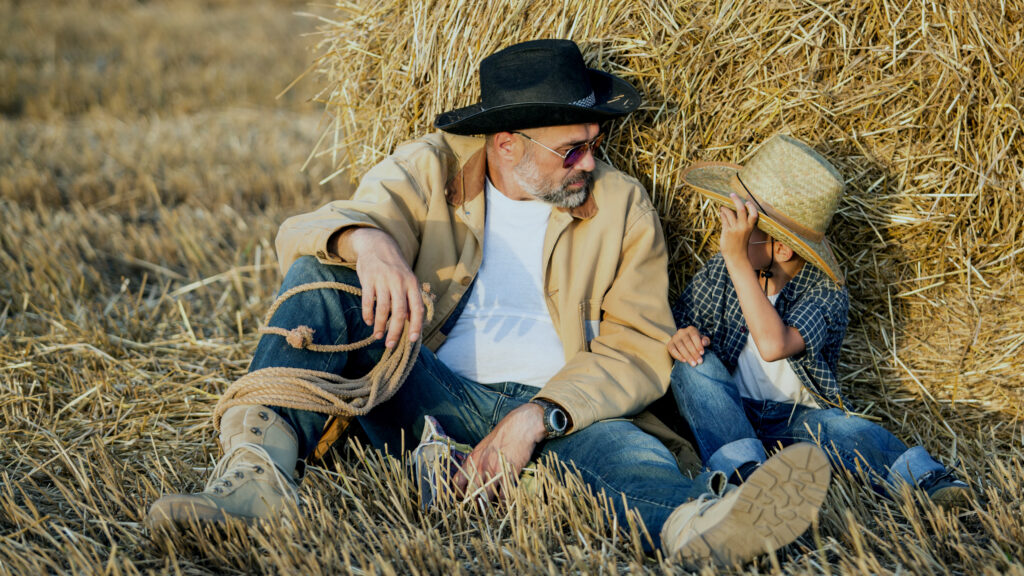 A man wearing glasses, a black hat, and casual clothes sits on the ground leaning against a hay bale, holding a rope. Beside him, a child in a straw hat, checked shirt, and jeans also sits, partially hiding their face with the hat. Both are in a harvested field.