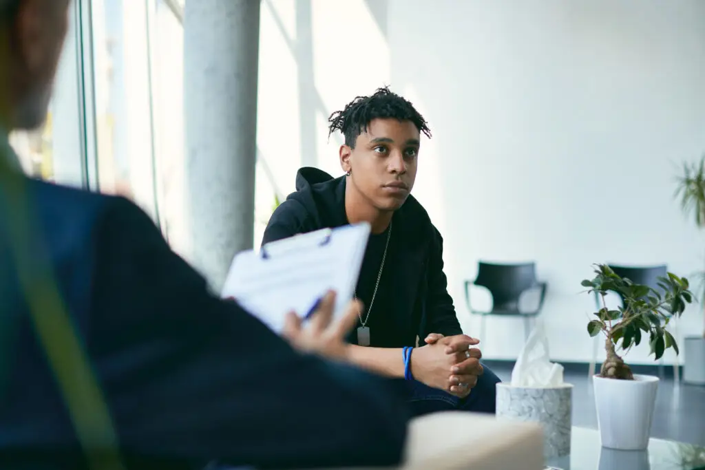 A young person with short, curly hair sits attentively on a couch, facing an unseen person holding a clipboard. The background includes large windows, a concrete pillar, and a small plant on a table next to a box of tissues.