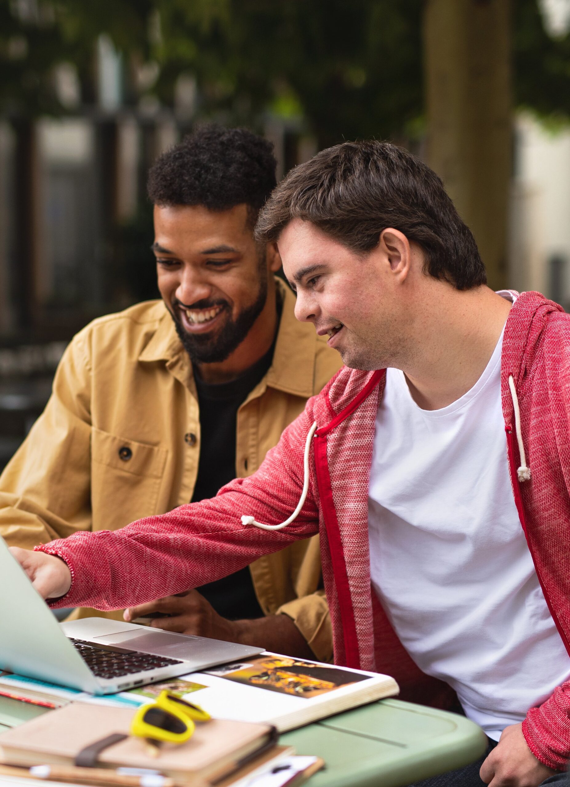 Two people are sitting at a table outdoors, looking at a laptop. The person on the left is wearing a tan jacket and has a beard, while the person on the right is in a red hoodie, pointing at the screen. There are notebooks, a book, and yellow sunglasses on the table.