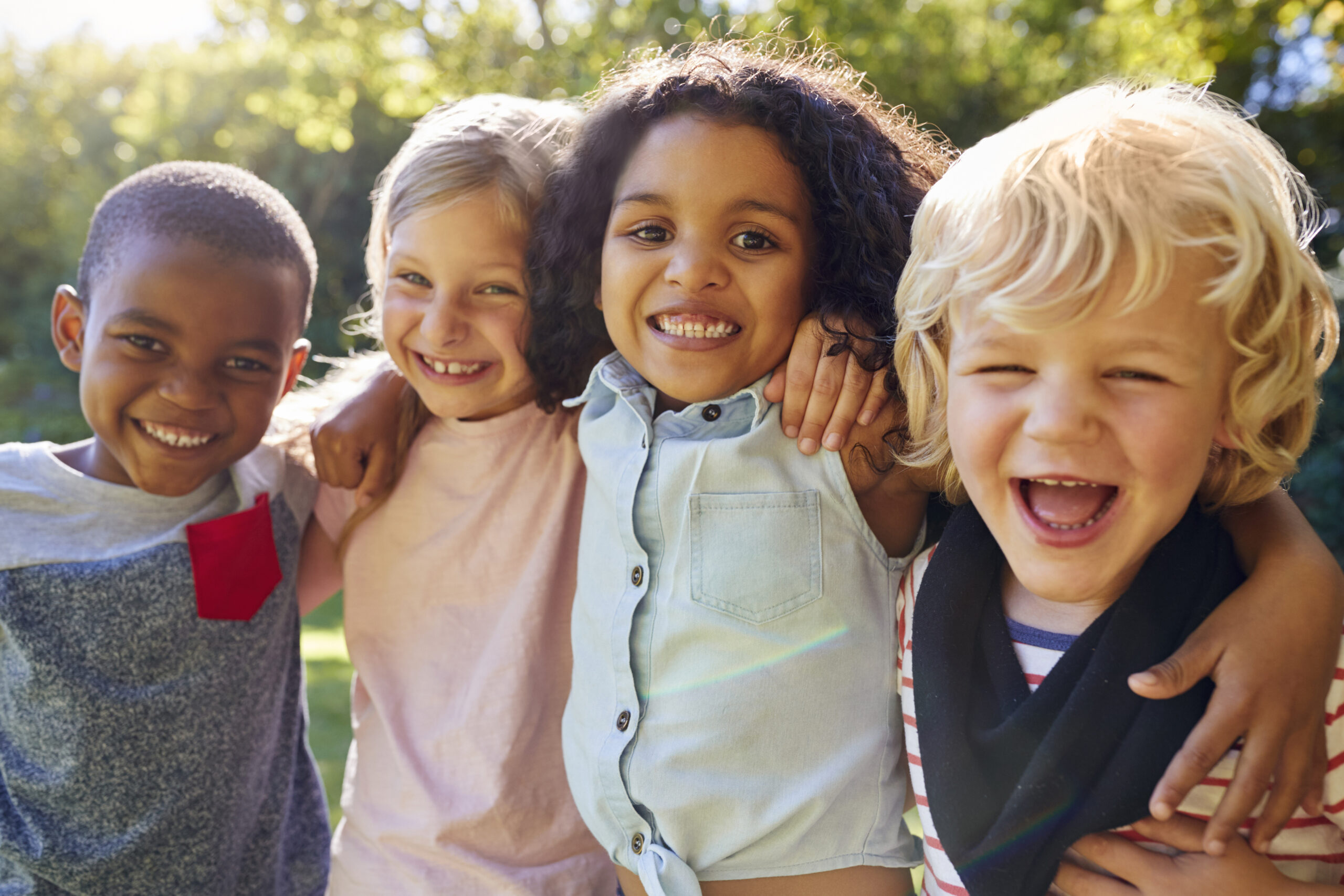 Four children stand outdoors with their arms around each other's shoulders, smiling and laughing. The background is a green, sunlit park with trees. The children show a diverse range of appearances.