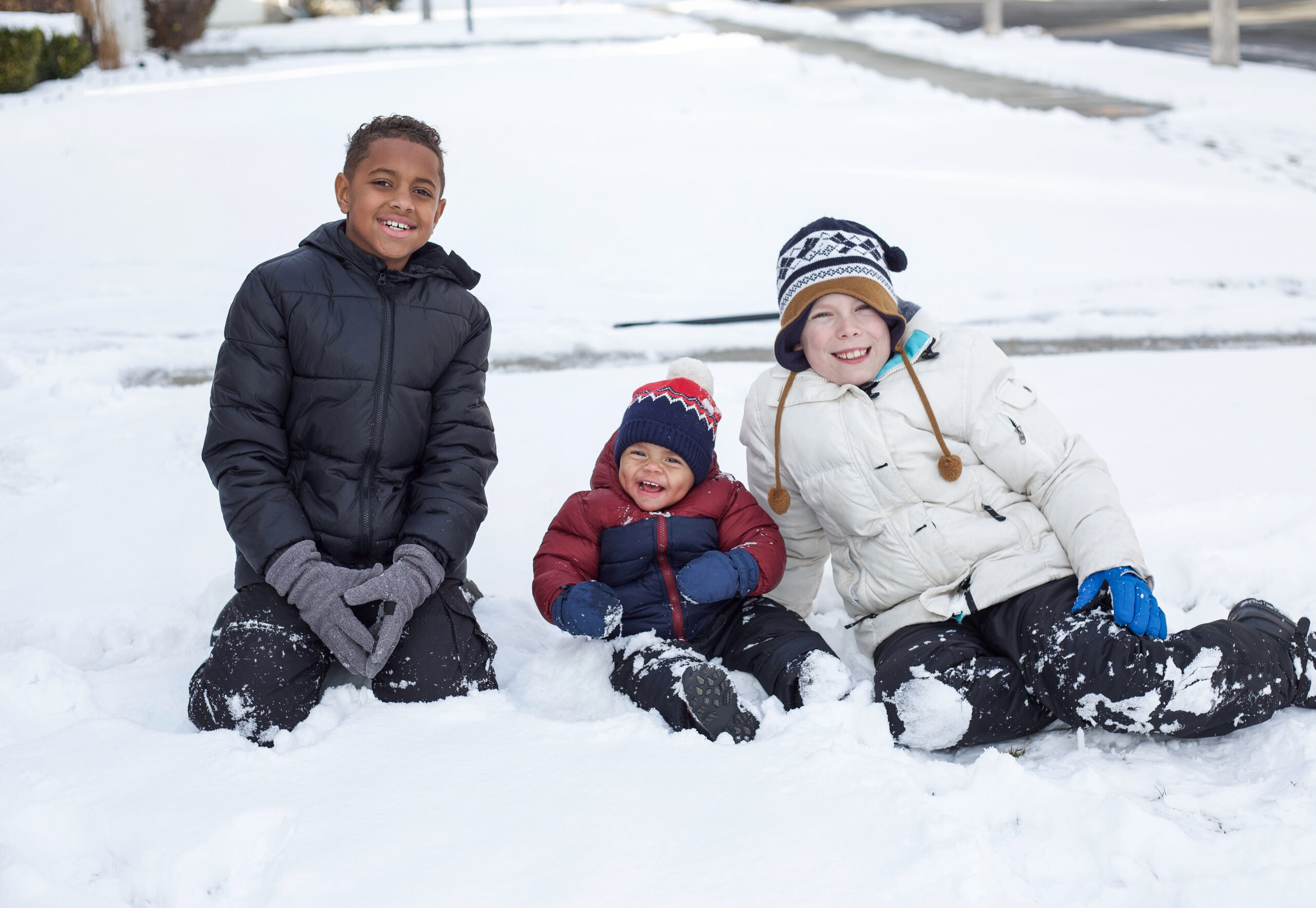 Three children are sitting in the snow. The child on the left is wearing a black jacket and gray gloves. The child in the middle is wearing a red jacket and a blue hat with a red pom-pom. The child on the right is wearing a white jacket and a knit hat.