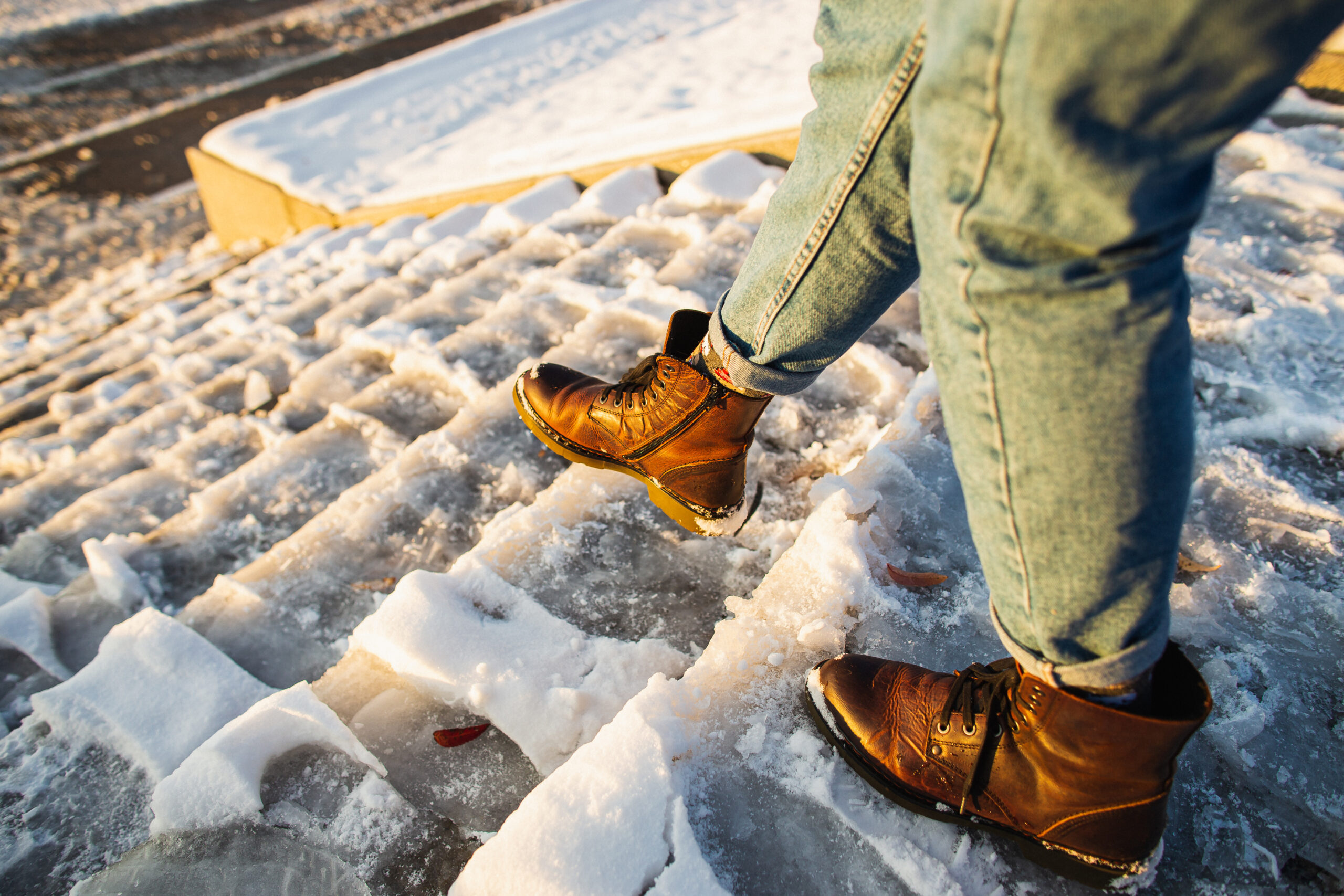 A person in blue jeans and brown boots walks up icy, snow-covered steps. The image captures their legs and feet, highlighting the texture of both the boots and the ice.