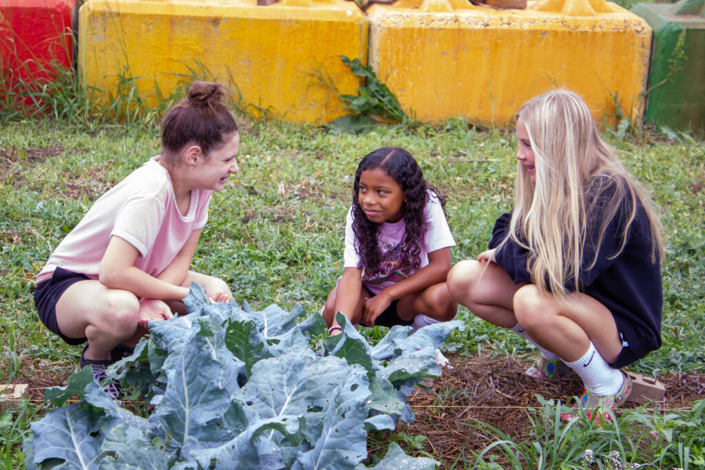 Three girls are outdoors in a garden, looking down at a large leafy plant. The girl on the left has a high bun and is smiling. The middle girl has long curly hair, and the girl on the right has long blonde hair and is wearing a black sweatshirt.