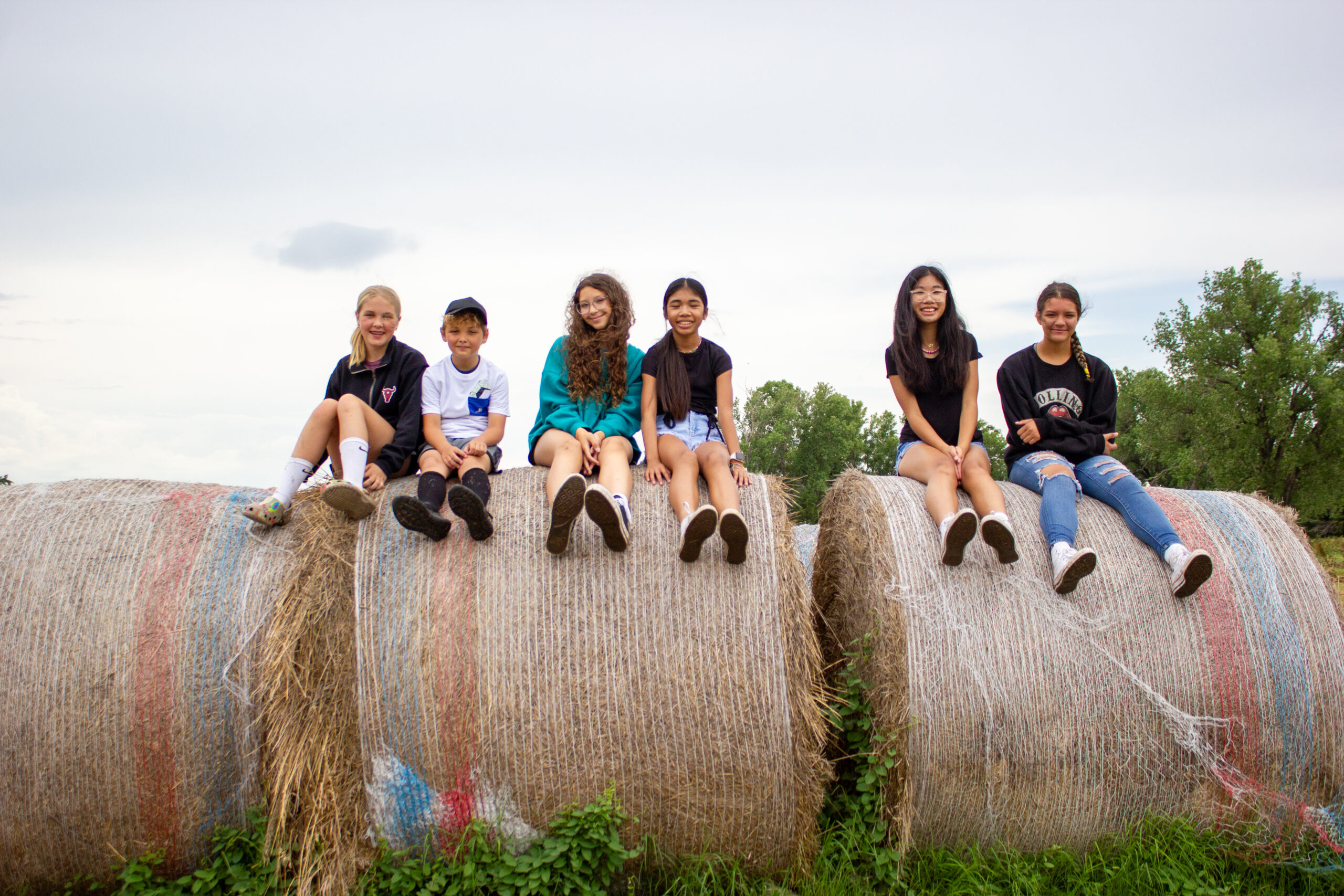 A group of six people, four girls and two boys, sit on large hay bales outdoors. They are casually dressed, wearing shorts and t-shirts. The background features a cloudy sky and green trees.