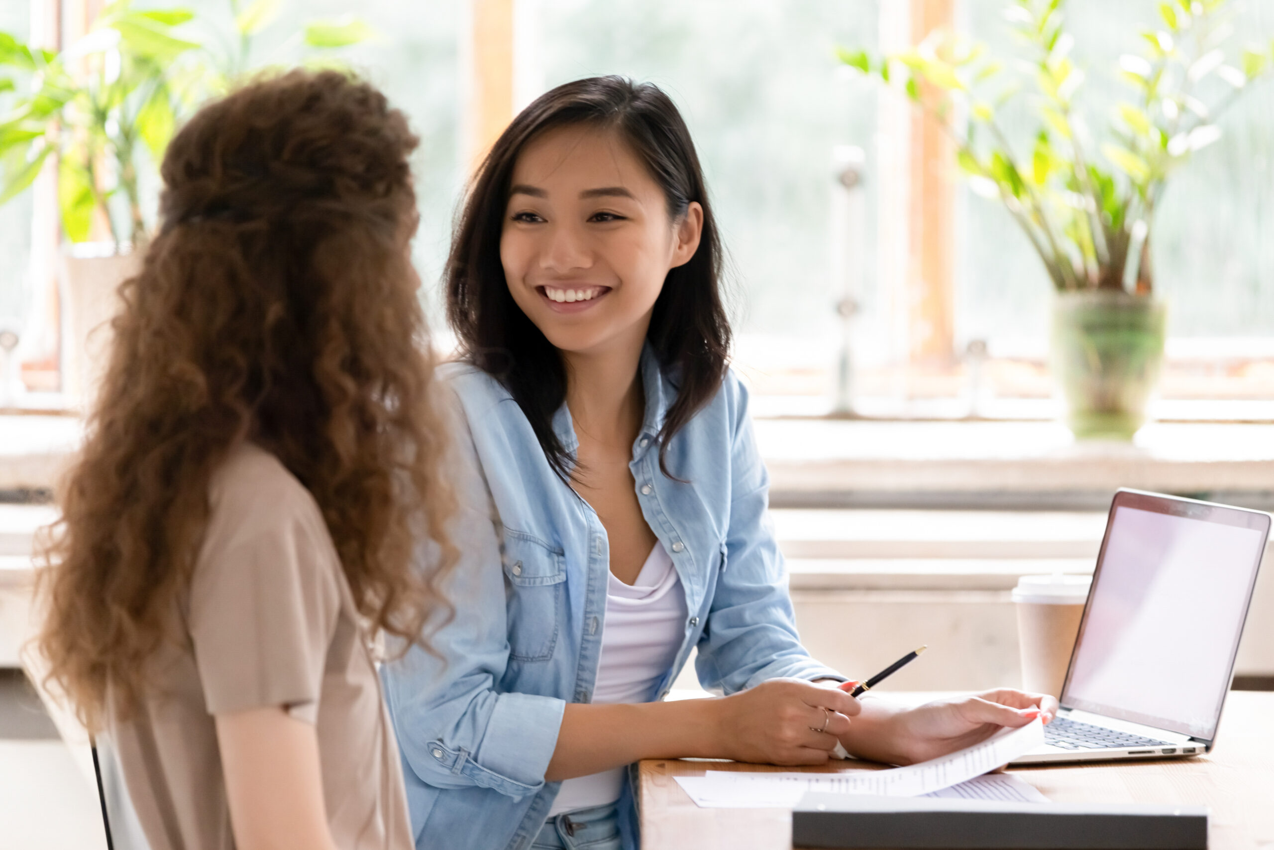 Two women are sitting at a wooden table, engaged in a conversation. One woman, holding a pen, has a notebook and a laptop in front of her. They are indoors, with green plants and a large window in the background. Both women are smiling.