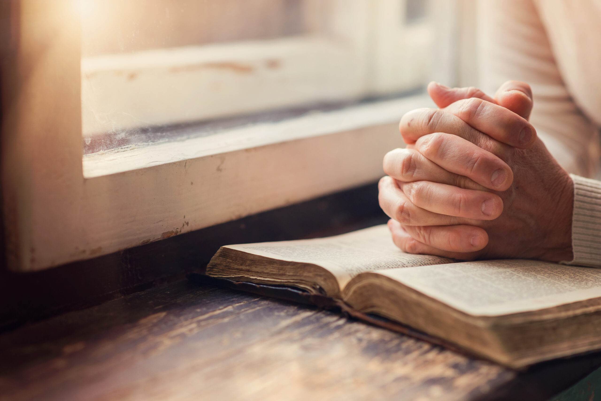 Person with clasped hands resting on an open book near a window, with sunlight streaming in. The scene suggests a moment of reflection or prayer. The window and table have a rustic appearance.