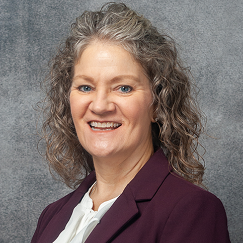 A woman with curly, shoulder-length gray hair wearing a maroon blazer and white blouse smiles against a gray textured background.