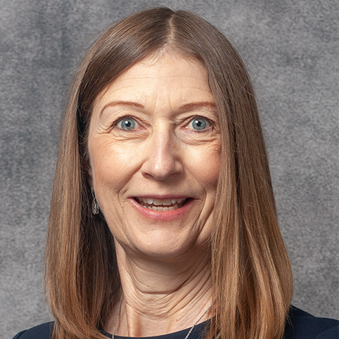 A woman with straight, light brown hair is smiling at the camera. She has blue eyes and is wearing a dark top. The background is a plain gray color.