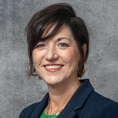 A person with short dark hair and wearing gold hoop earrings is smiling at the camera. They are dressed in a navy blue blazer over a green top. The background is a plain gray textured surface.