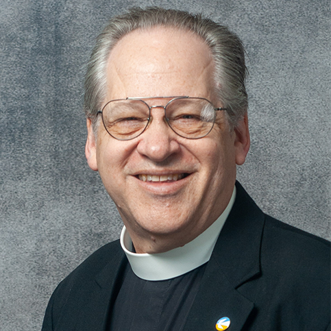 A man with gray hair and glasses is smiling. He is wearing a clerical collar, a black jacket, and a pin on his lapel. The background is gray.