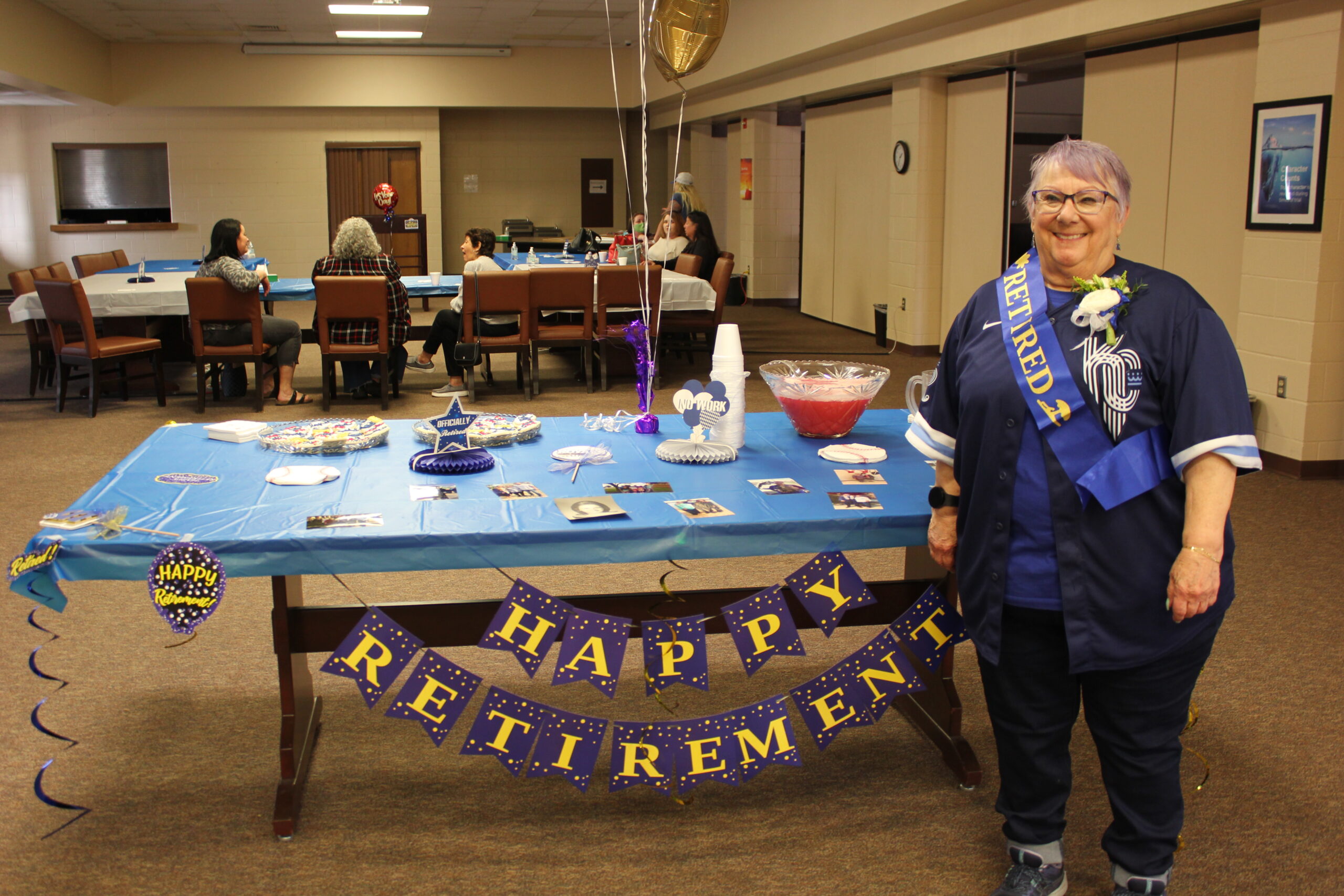 A woman in a "Retired" sash and shirt stands in front of a decorated table at a retirement party. The table has a blue tablecloth, snacks, a punch bowl, party decorations, and a "Happy Retirement" banner. People are seated in the background.