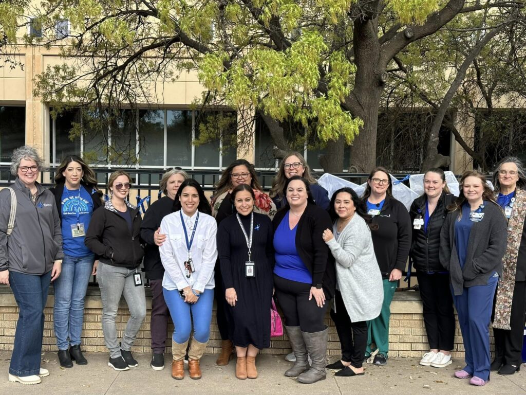 A group of fifteen people stands outside in front of a building and tree. They are dressed in casual and professional attire, with some wearing name badges. They are smiling and posing for the photo.
