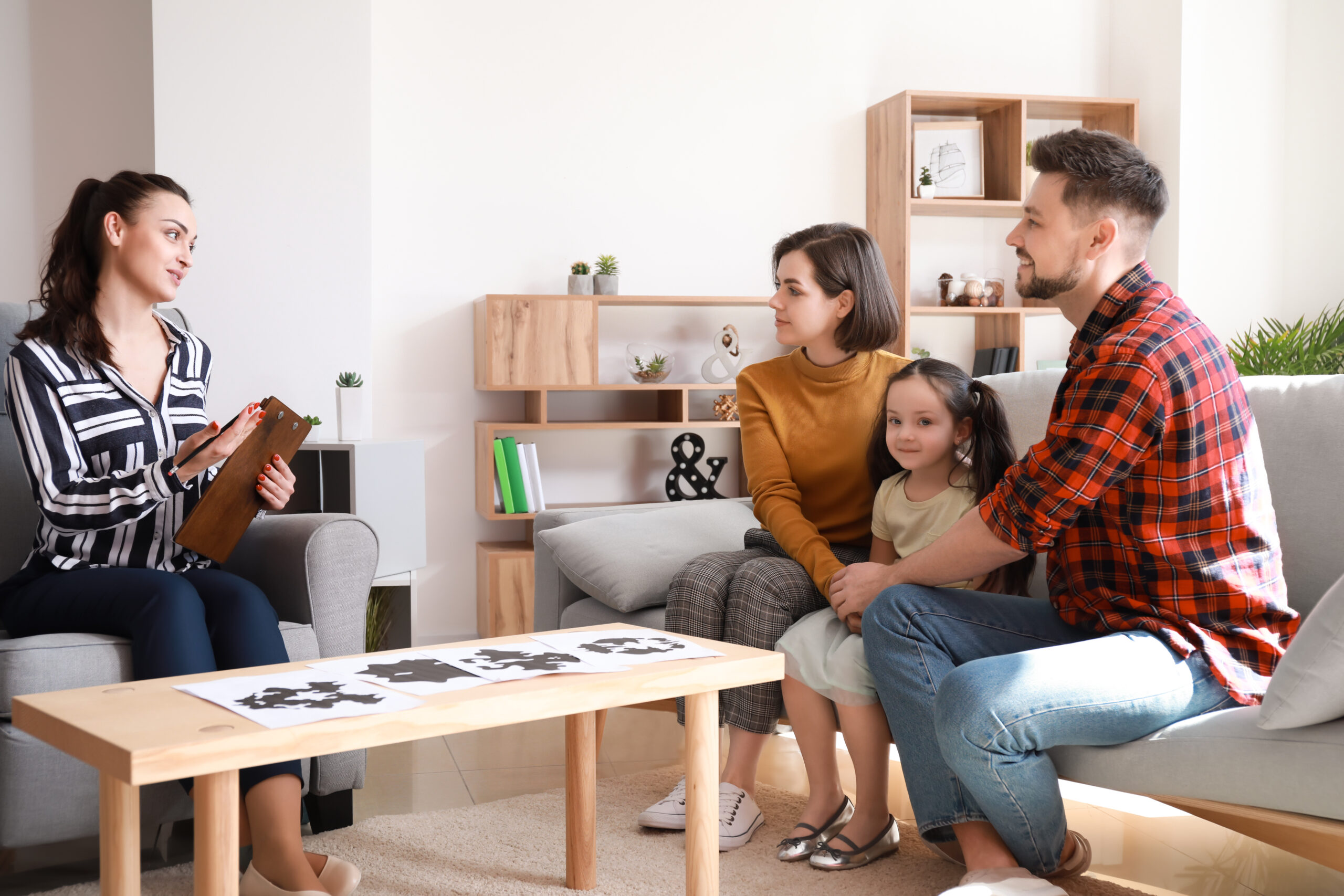 A woman holding a clipboard sits on a chair and speaks to a family of three seated on a couch. The family includes a man, a woman, and a young girl. They are in a living room with wooden shelves, books, and plants in the background.