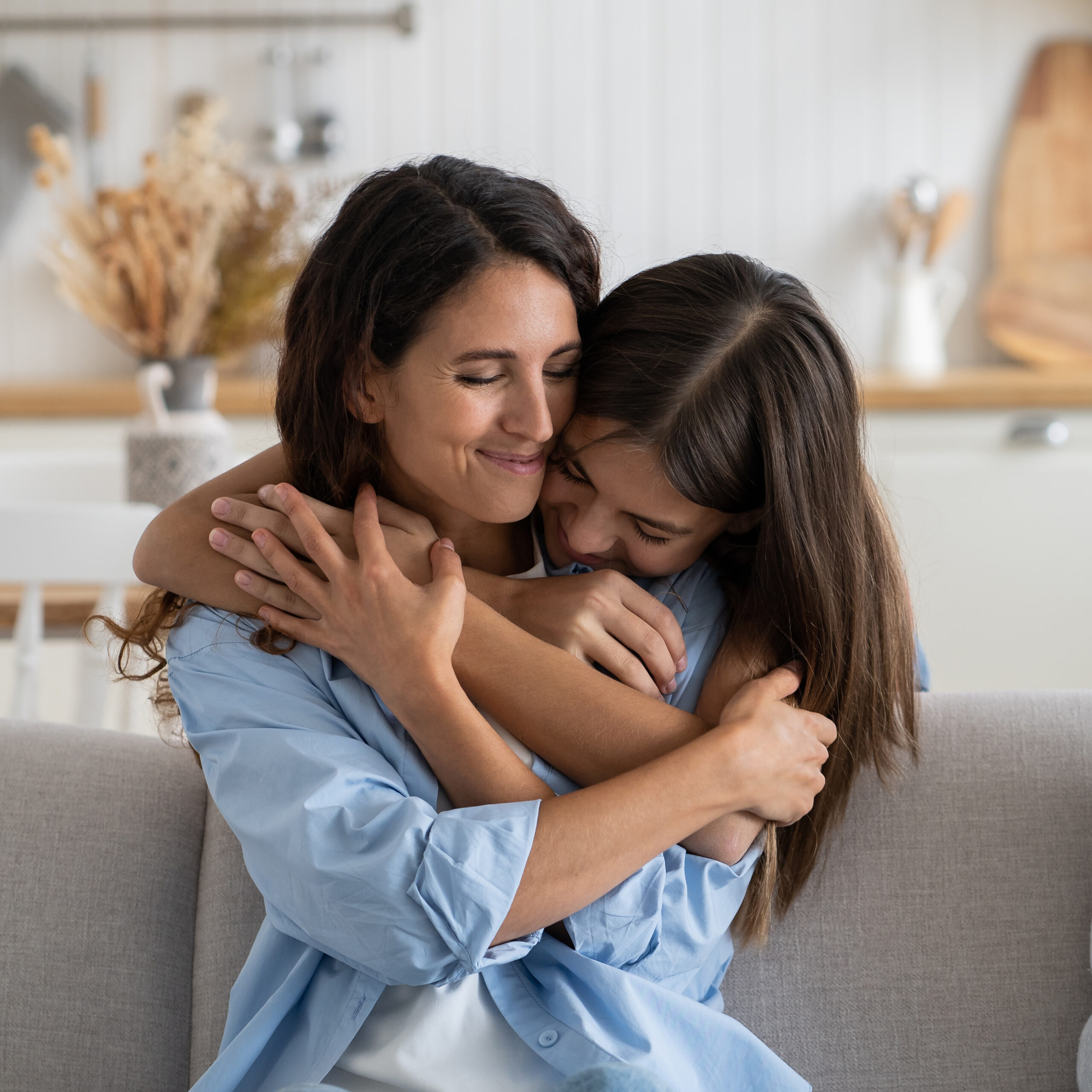 Two women are sitting on a sofa in a cozy living room, embracing each other. Both have long brown hair and are wearing light blue shirts. The background features kitchen elements like wooden utensils and dried plants.