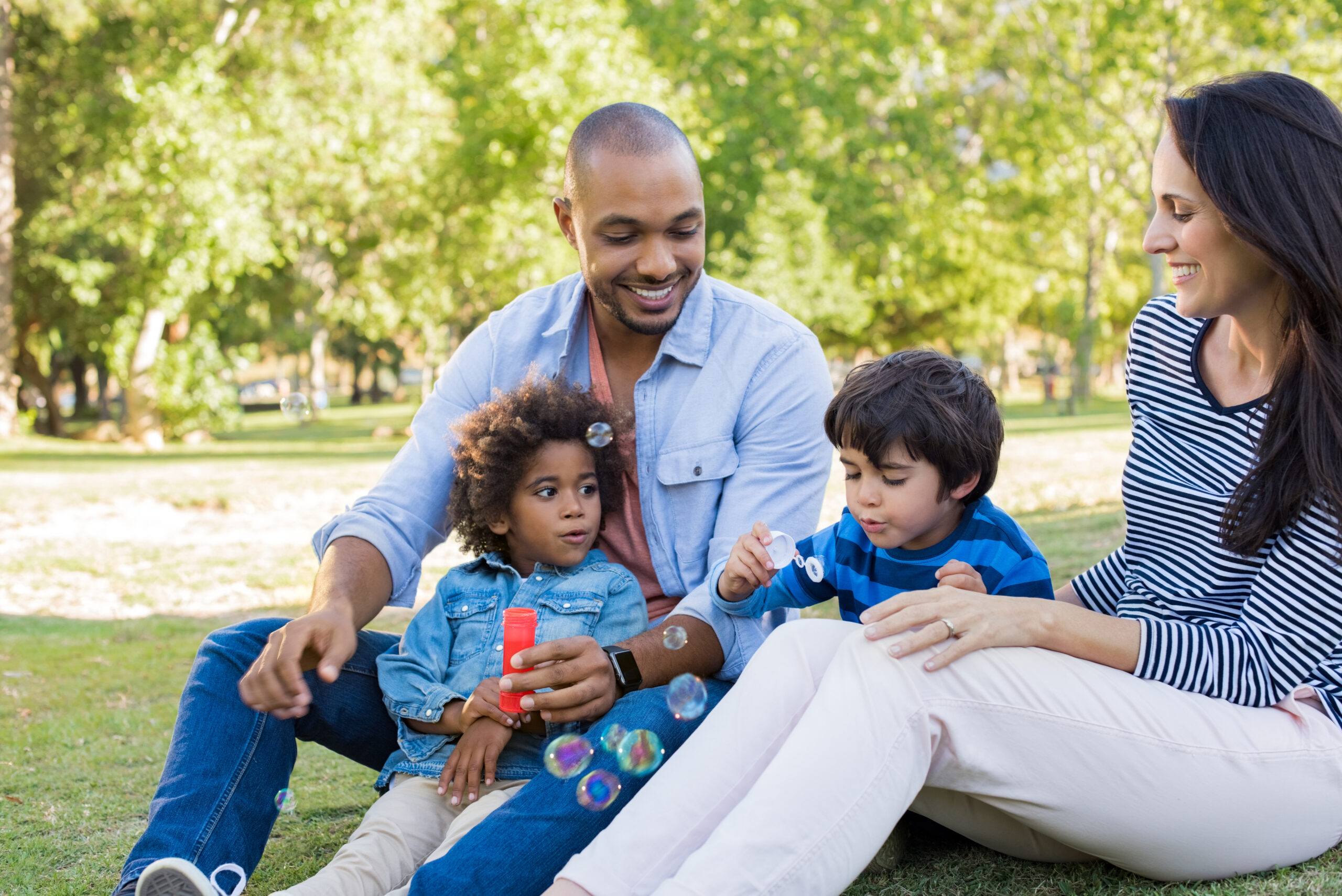 A group of four people are sitting on grass in a park on a sunny day. A man and woman are watching two children blowing bubbles. The man is holding a young child on his lap, while the older child is focused on blowing bubbles from a wand. Trees are in the background.