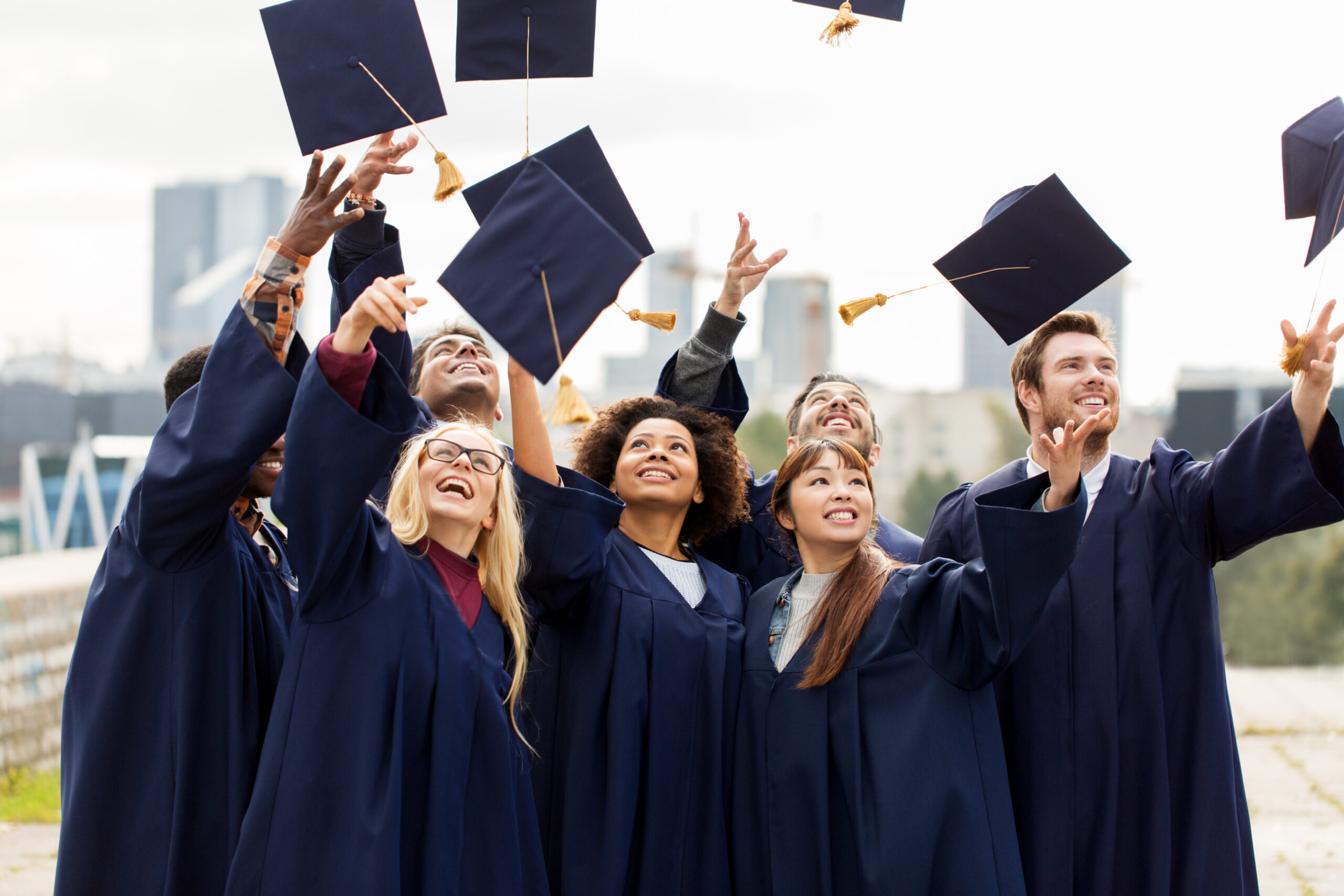 A group of graduates in blue gowns and caps celebrate outdoors by tossing their caps into the air. The background includes tall city buildings and a cloudy sky. The graduates appear joyful, with hands raised towards the caps.