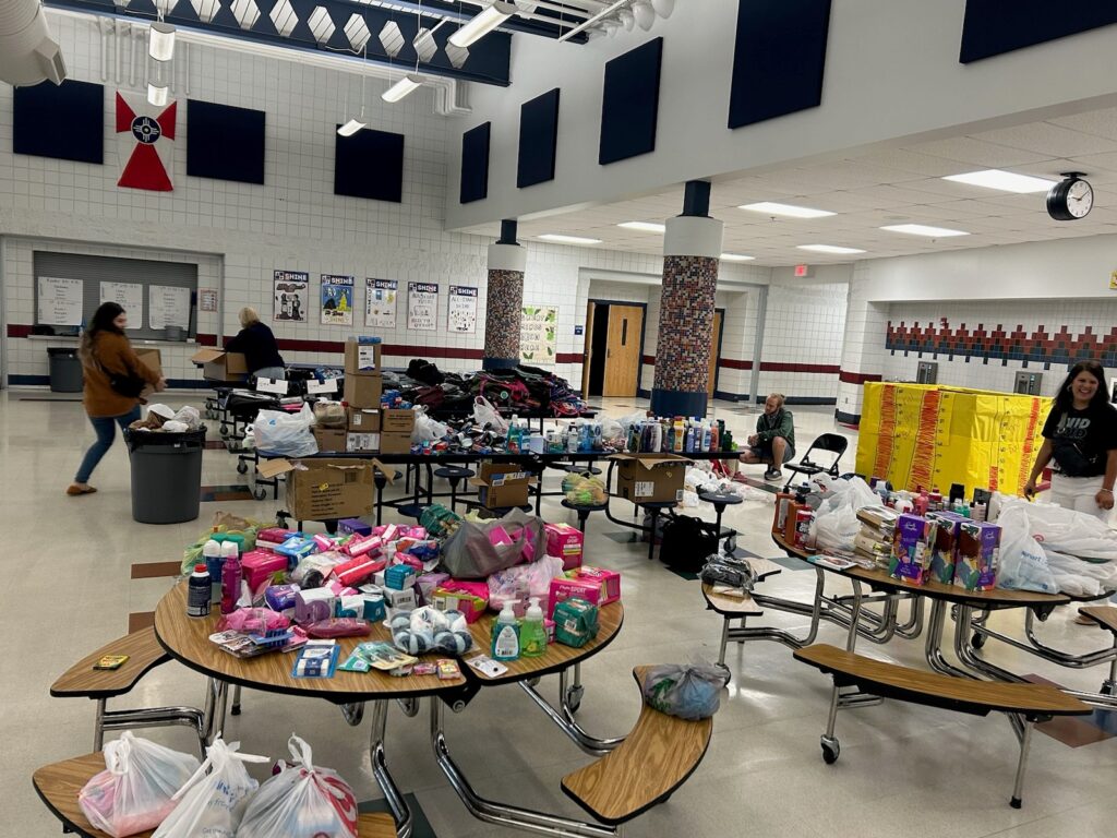 A school cafeteria is filled with various donation items, including toiletries, cleaning supplies, clothes, and canned goods. Several tables are covered with items, and a few volunteers are organizing and sorting the donations.