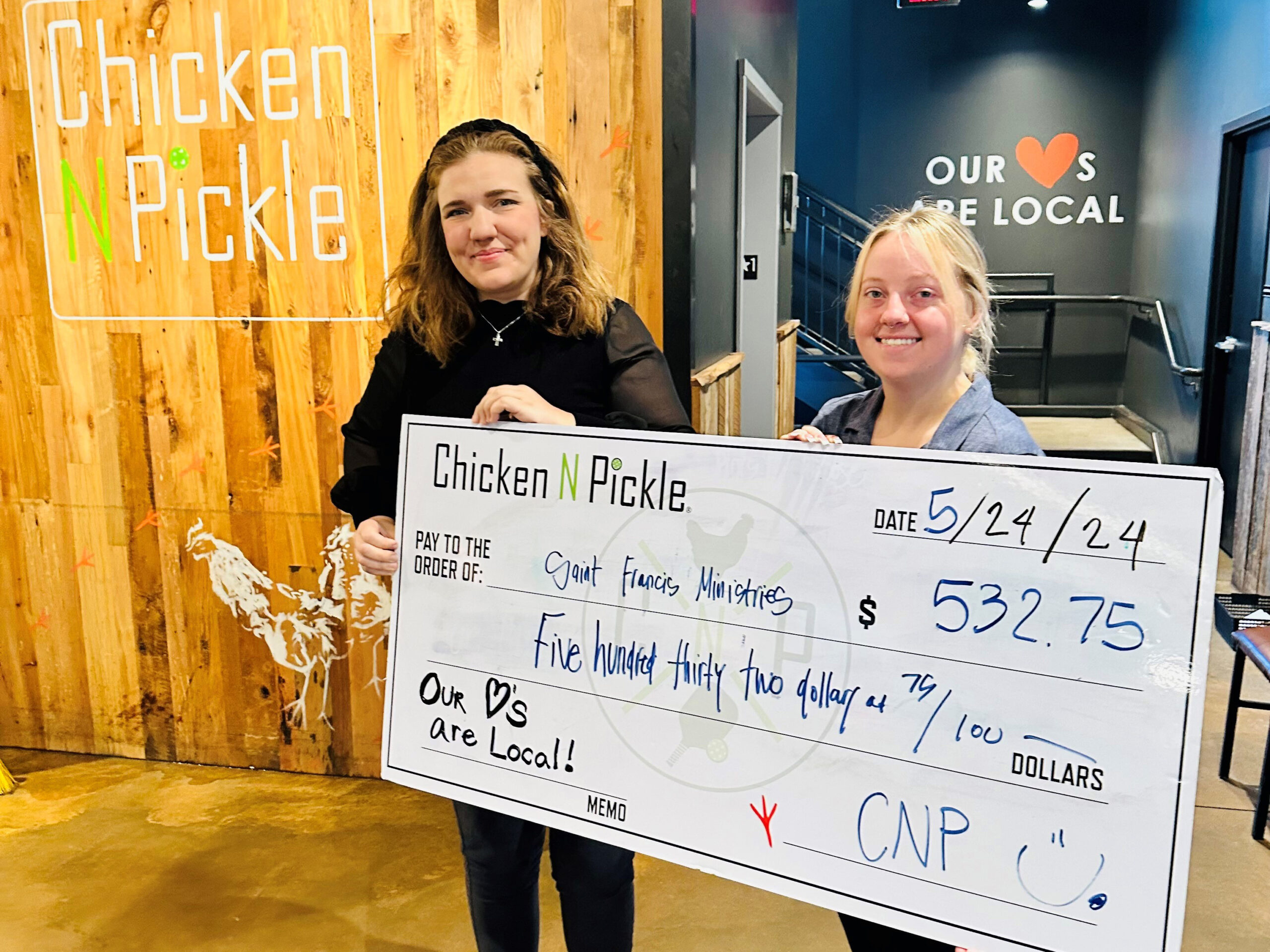 Two individuals smile while holding a large check worth $532.75 made out to Saint Francis Ministries from Chicken N Pickle. The presentation date is 5/24/24. They are standing in front of a wooden wall with the Chicken N Pickle logo and a heart symbol.