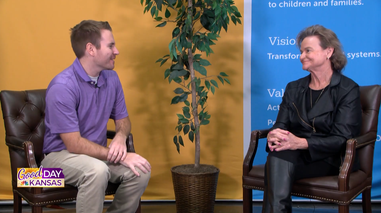 Two people sit in armchairs facing each other on a TV show set with "Good Day Kansas" branding. A potted plant is between them. The left person wears a purple shirt and gray pants, and the right person is in a black outfit. A blue background has text.