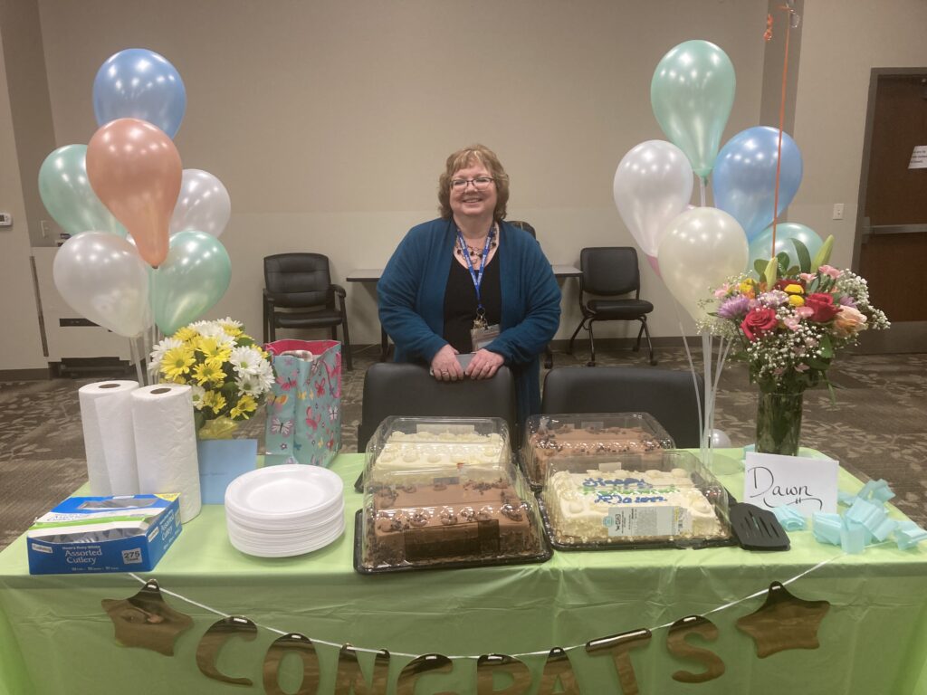 A woman stands behind a table covered with a green cloth and decorated with a "Congrats" banner. The table has cakes, paper plates, wrapped gifts, a bouquet of flowers, and balloons. The woman is smiling, wearing a lanyard, and has curly hair and glasses.