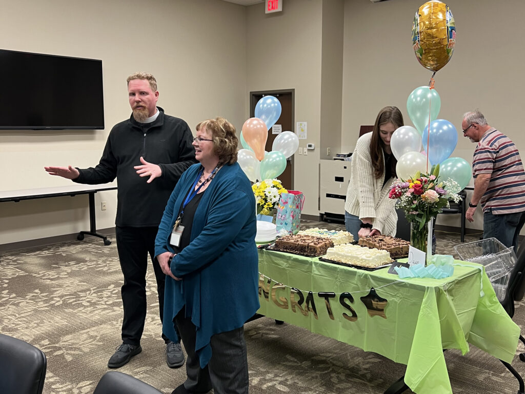 A man and a woman stand in front of a table with cakes and a "Congrats" sign. The man is speaking while the woman smiles. Another woman is cutting cake, and a man in striped shirt is in the background. Balloons and flowers decorate the table.