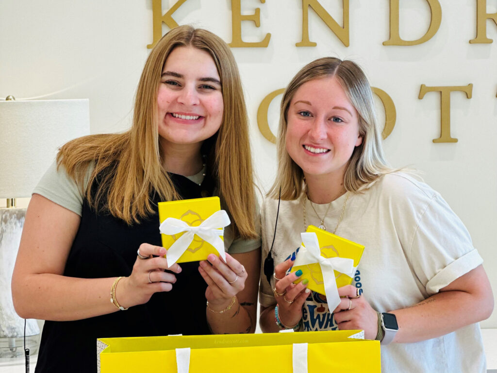Two smiling women holding yellow gift boxes with white ribbons. They are standing in front of a wall with partially visible text in the background. One woman is in a black shirt, and the other is in a white shirt. There is a yellow shopping bag in front of them.