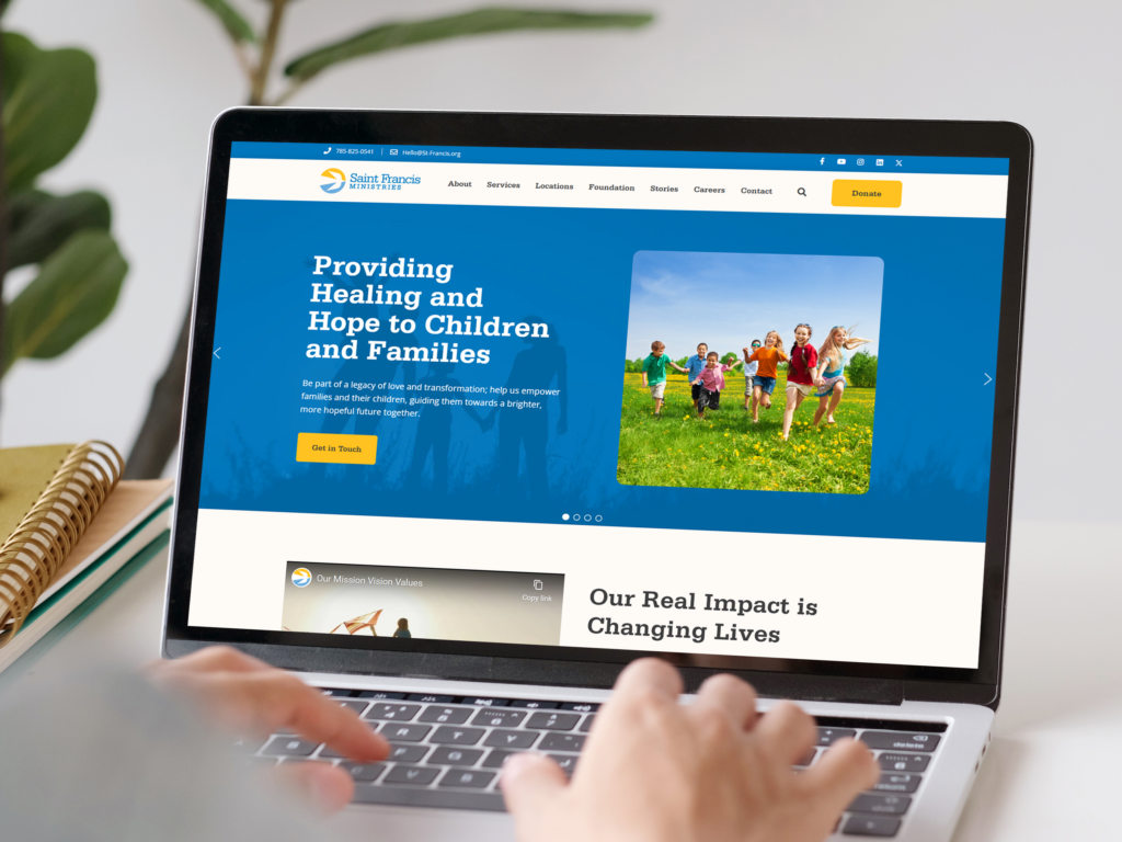 A person is using a laptop, displaying a website for a children's health organization. The website features a header with the text "Providing Healing and Hope to Children and Families" next to an image of children running in a field.