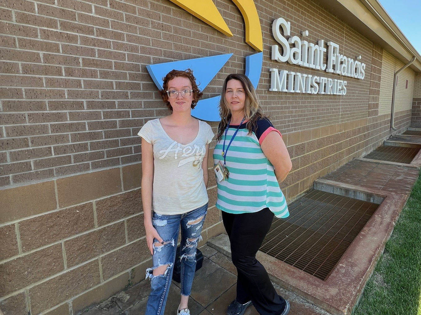 Two women stand in front of a brick building with a sign that reads "Saint Francis Ministries." One woman with glasses and curly hair is wearing a light t-shirt and ripped jeans. The other woman has long hair and is wearing a striped shirt with a name badge.