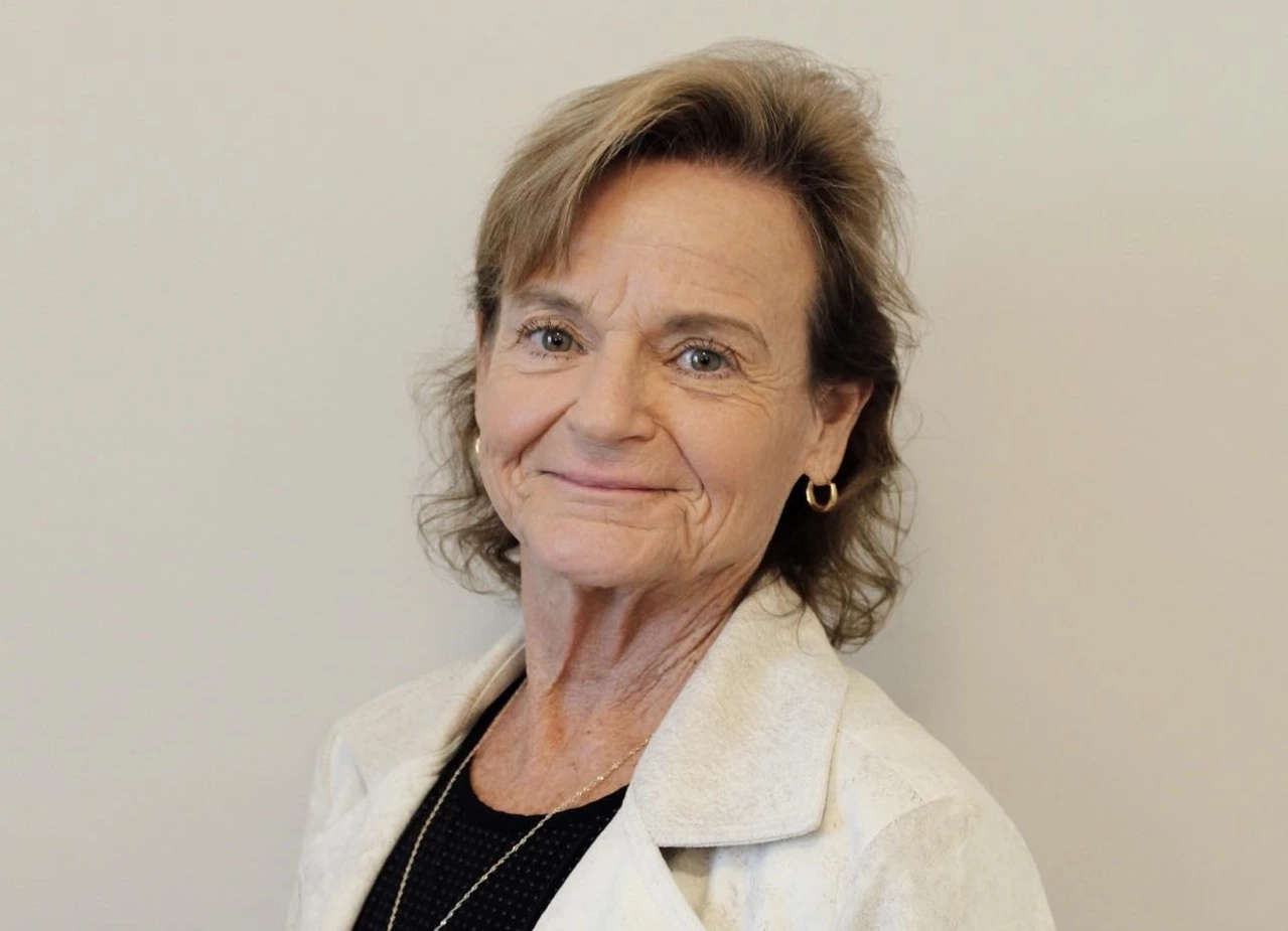 A woman with light brown hair and wearing a white jacket and black shirt is smiling slightly. She is standing against a plain beige background. She has gold hoop earrings and a necklace.