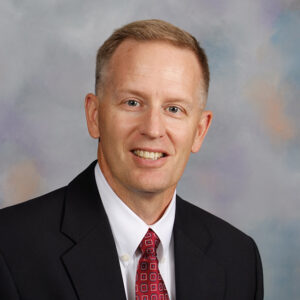 A middle-aged man with short, light brown hair is wearing a navy suit, white shirt, and red patterned tie. He is smiling and posing against a backdrop with a blend of grey, blue, and peach colors.