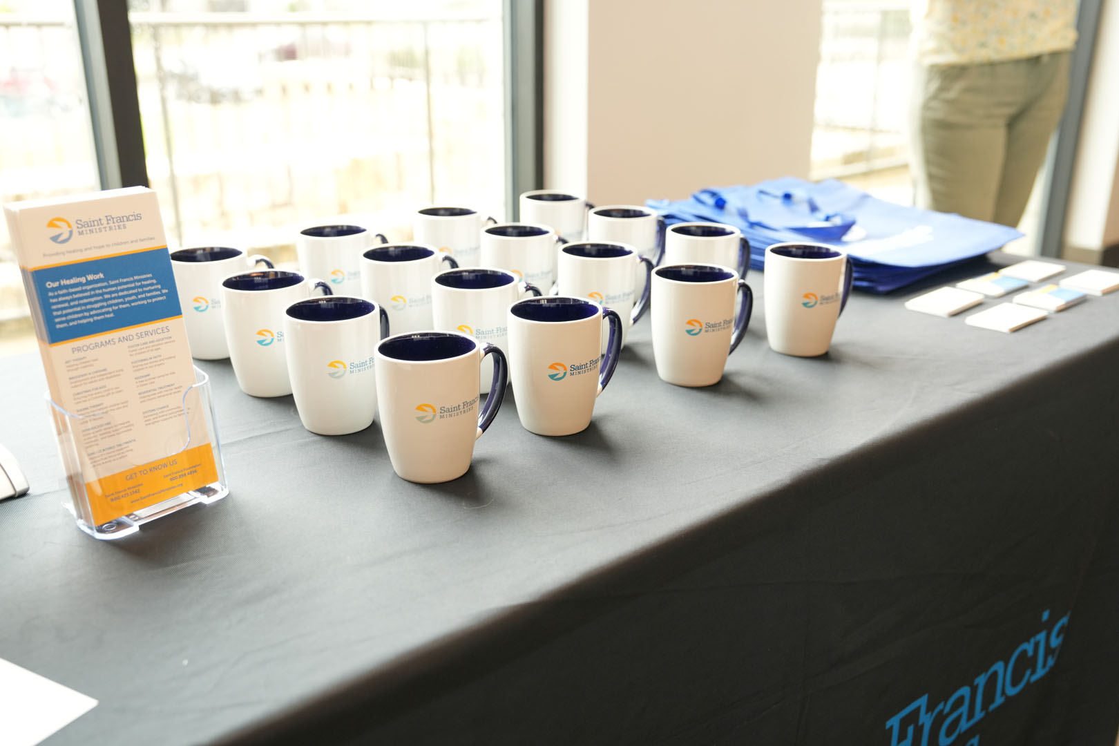 A table features white mugs with blue interiors and a logo, arranged in rows. On the left is a brochure display with a brochure about services and a logo. Blue tote bags are stacked to the right. The tablecloth is black, with partially visible text.