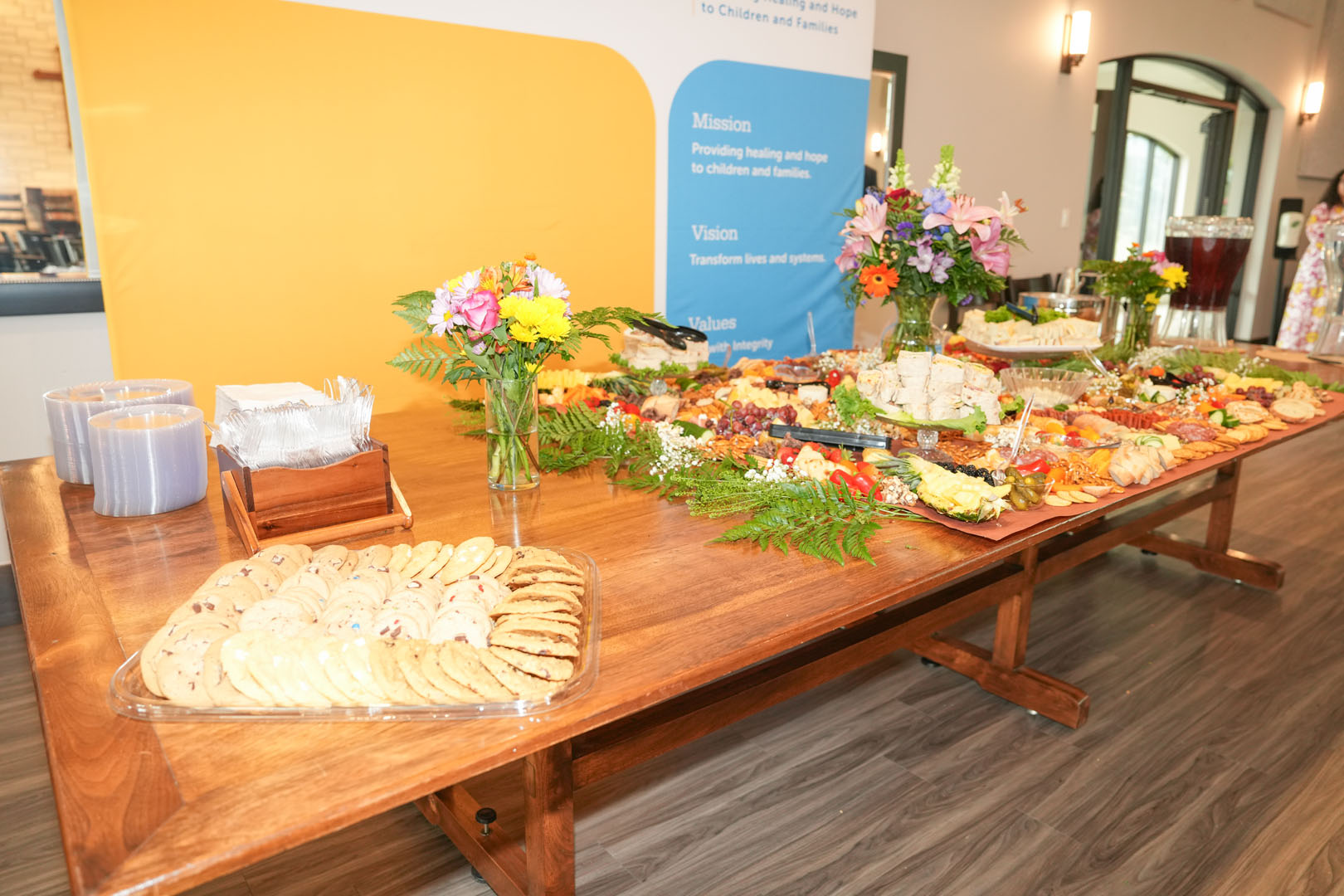 A wooden table is set up with an assortment of food for an event. Offerings include cookies, various cheeses, fruits, and other appetizers. There are flower arrangements and a background banner displaying text about the organization's mission and vision.