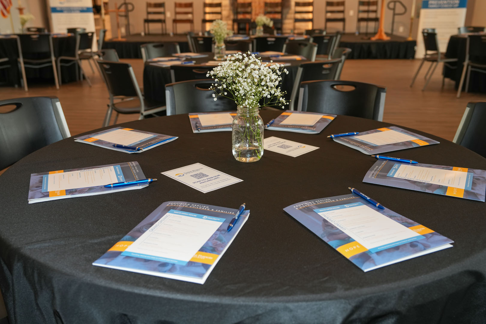 A round table with eight chairs is set up with conference materials, including booklets, pens, and informational sheets arranged at each seat. A vase with small white flowers is placed in the center of the table. Other tables and chairs are visible in the background.