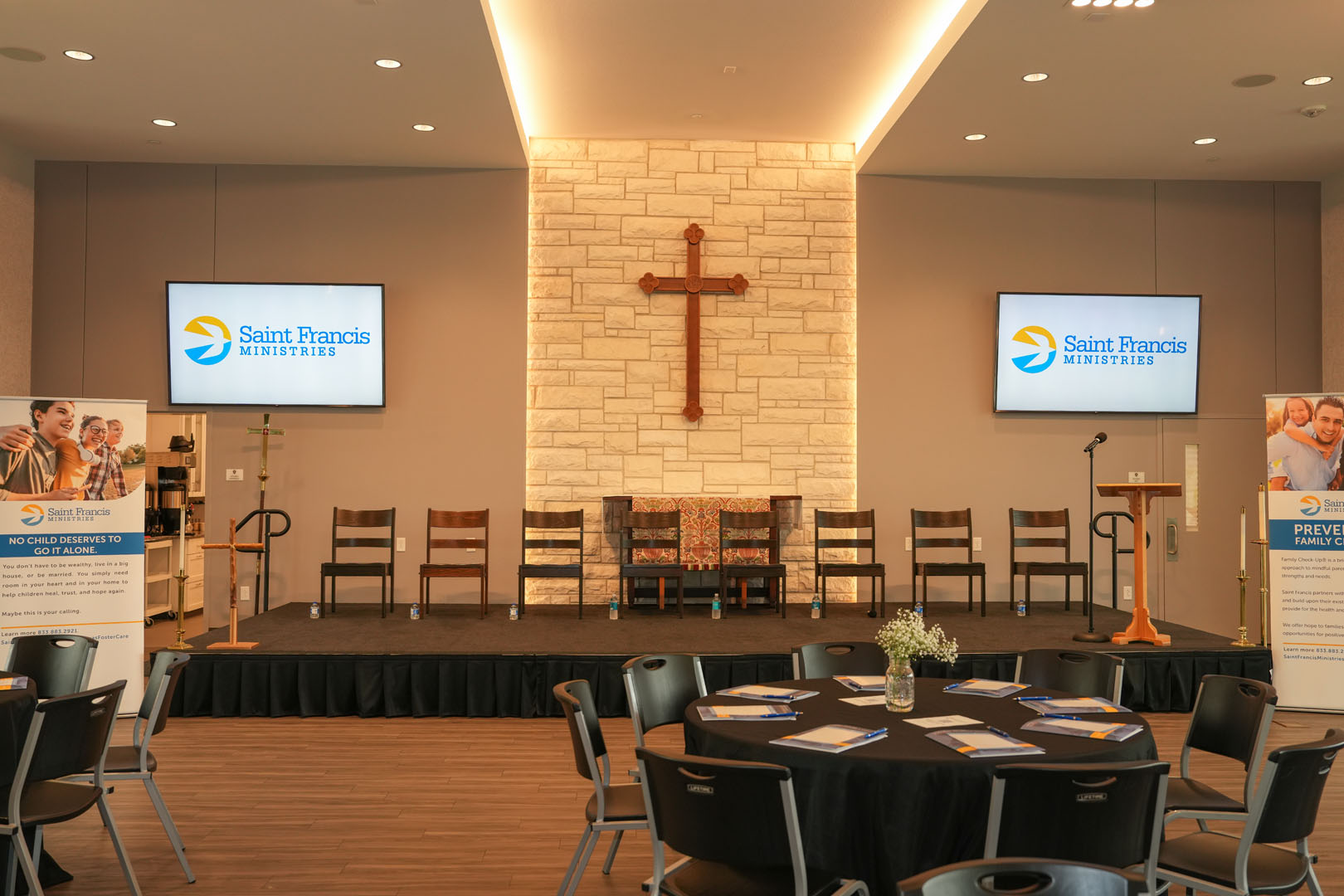 A stage in a church setting with six wooden chairs and two screens displaying the "Saint Francis Ministries" logo. A wooden cross is mounted on a brick wall behind the stage. The foreground shows several round tables with brochures and flower arrangements.