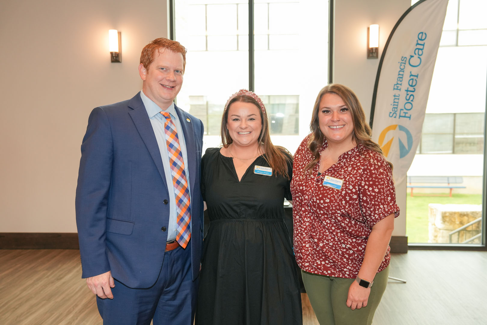 Three people stand indoors smiling at the camera. The man on the left is wearing a suit and tie, the woman in the middle is in a black dress, and the woman on the right is in a red patterned blouse and green pants. A "Saint Francis Foster Care" banner is visible in the background.