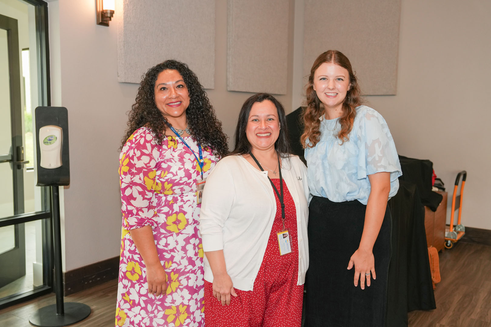 Three women stand together, all smiling at the camera. They are indoors, with a hand sanitizer dispenser on the left and a wheeled cart at the back right. The woman on the left wears a pink dress with floral patterns, the center woman wears a white cardigan over a red dress, and the woman on the right wears a light blue blouse with a black skirt.