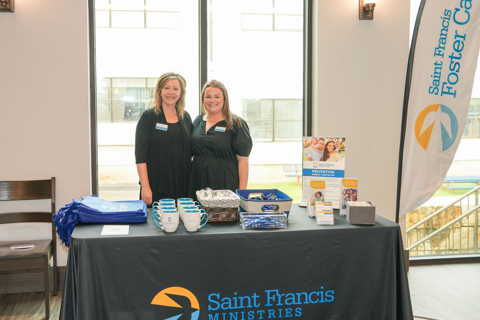 Two women stand behind a Saint Francis Ministries table at an event. The table displays promotional items, including mugs, brochures, and bags. A tall banner and window are in the background. Both women are smiling and wear black attire with name tags.