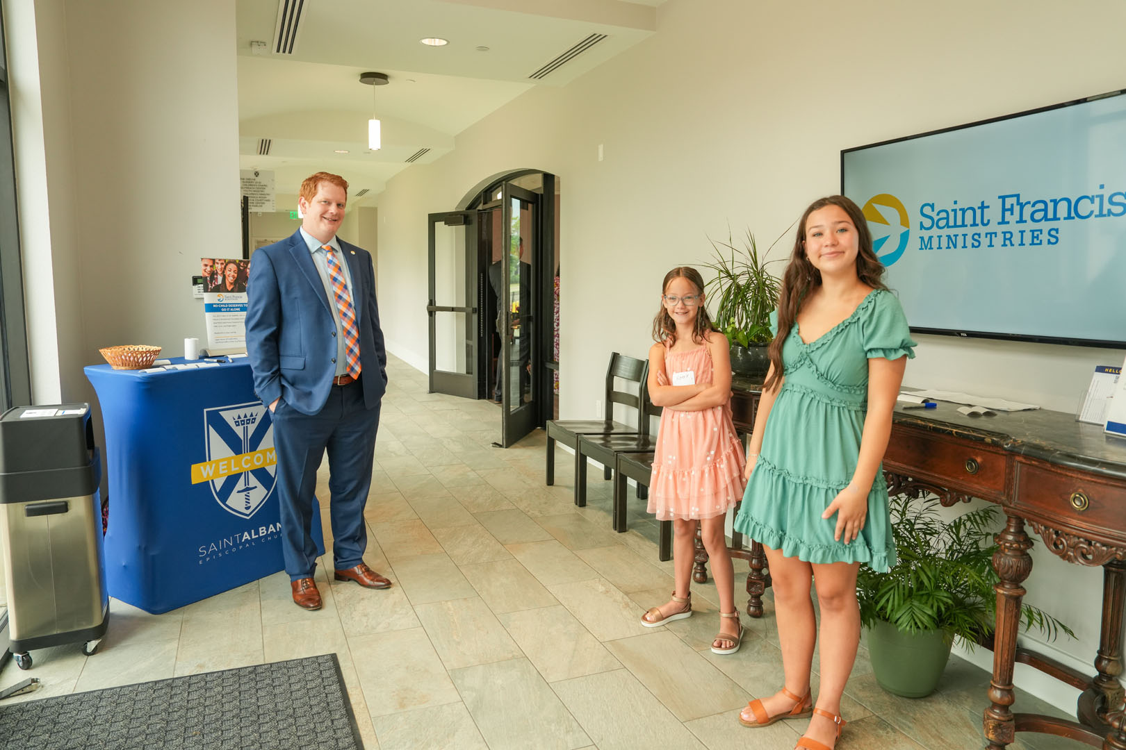 A man in a suit stands beside a welcome table with snacks and a Saint Francis Ministries banner. Two young girls in dresses stand near a wooden table with potted plants and a brochure holder. The setting appears to be an office or lobby area with a tiled floor.