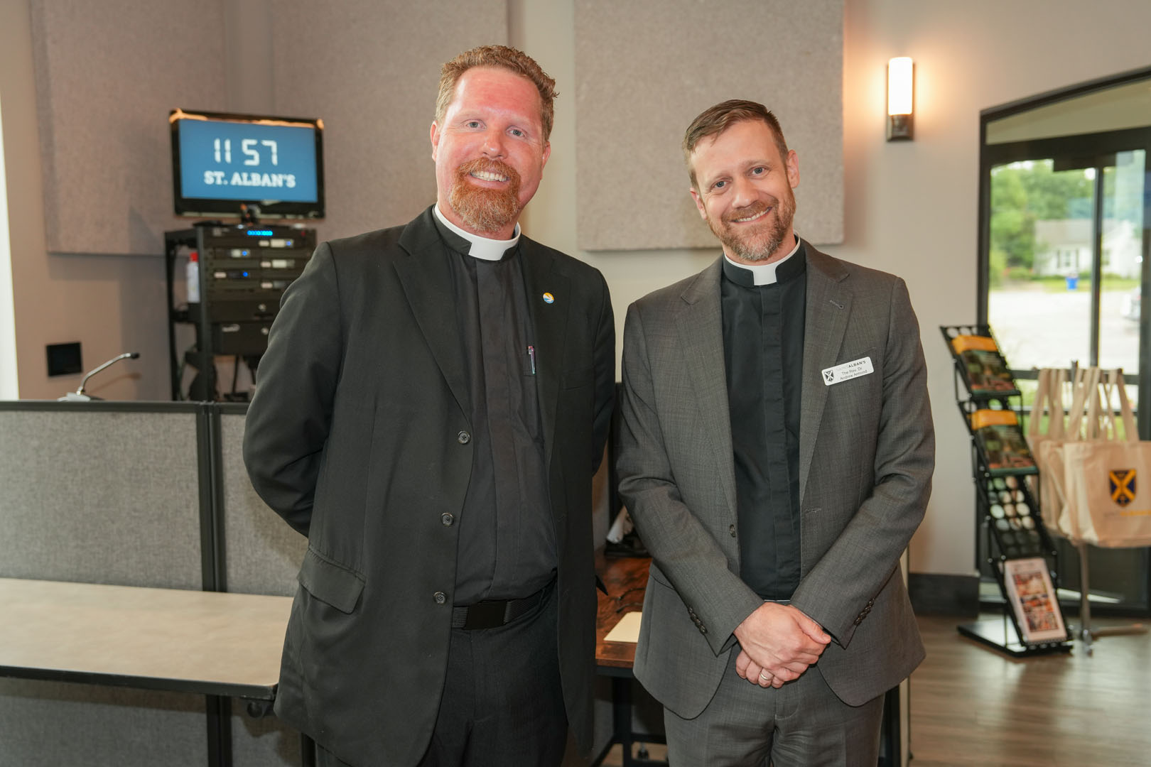 Two men dressed in clerical collars are standing side by side in a room with grey walls and soundproof panels. The man on the left wears a black suit and smiles at the camera. The man on the right, with a name tag, wears a grey suit and also smiles. A digital clock in the background shows "11:57" and the text "St. Alban's." Various items are displayed near the window on the right.