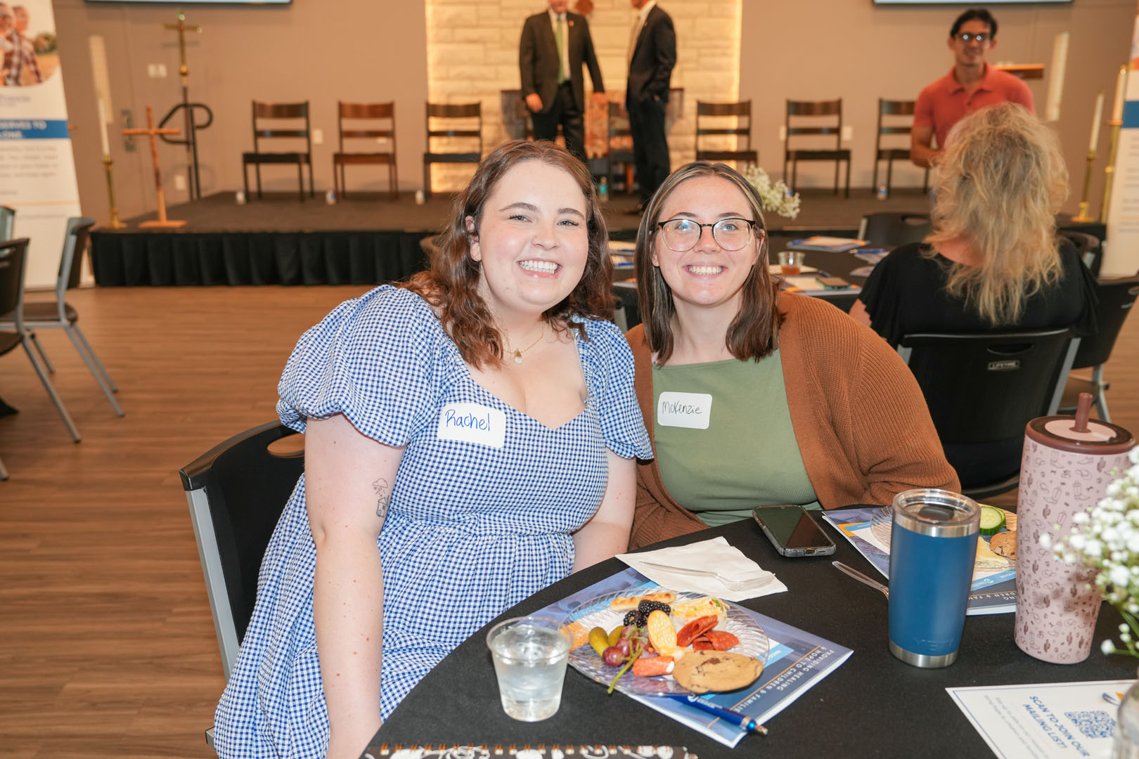 Two women are seated at a table, smiling at the camera. One is wearing a blue and white striped dress, and the other is in a brown cardigan over a green top. Food and drinks are on the table, and other people can be seen in the background.