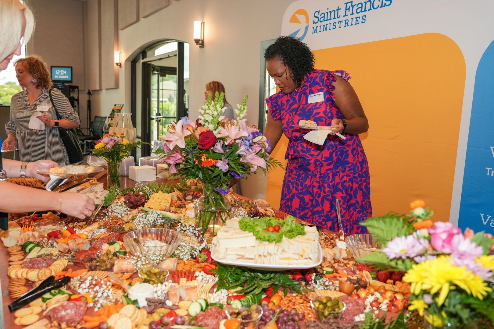 A woman in a floral-pattern dress serves herself from a large charcuterie table adorned with various cheeses, meats, fruits, and crackers. Other people are also serving themselves. A sign in the background reads "Saint Francis Ministries." The setting appears to be an indoor event.