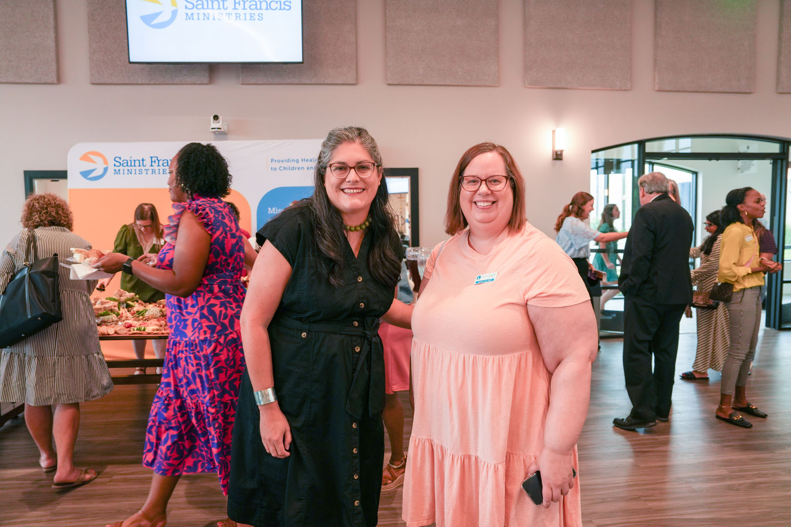 Two women are posing and smiling indoors at an event hosted by Saint Francis Ministries. The woman on the left wears a black dress, and the woman on the right wears a peach-colored dress. Other attendees are mingling in the background, and a food table is visible.