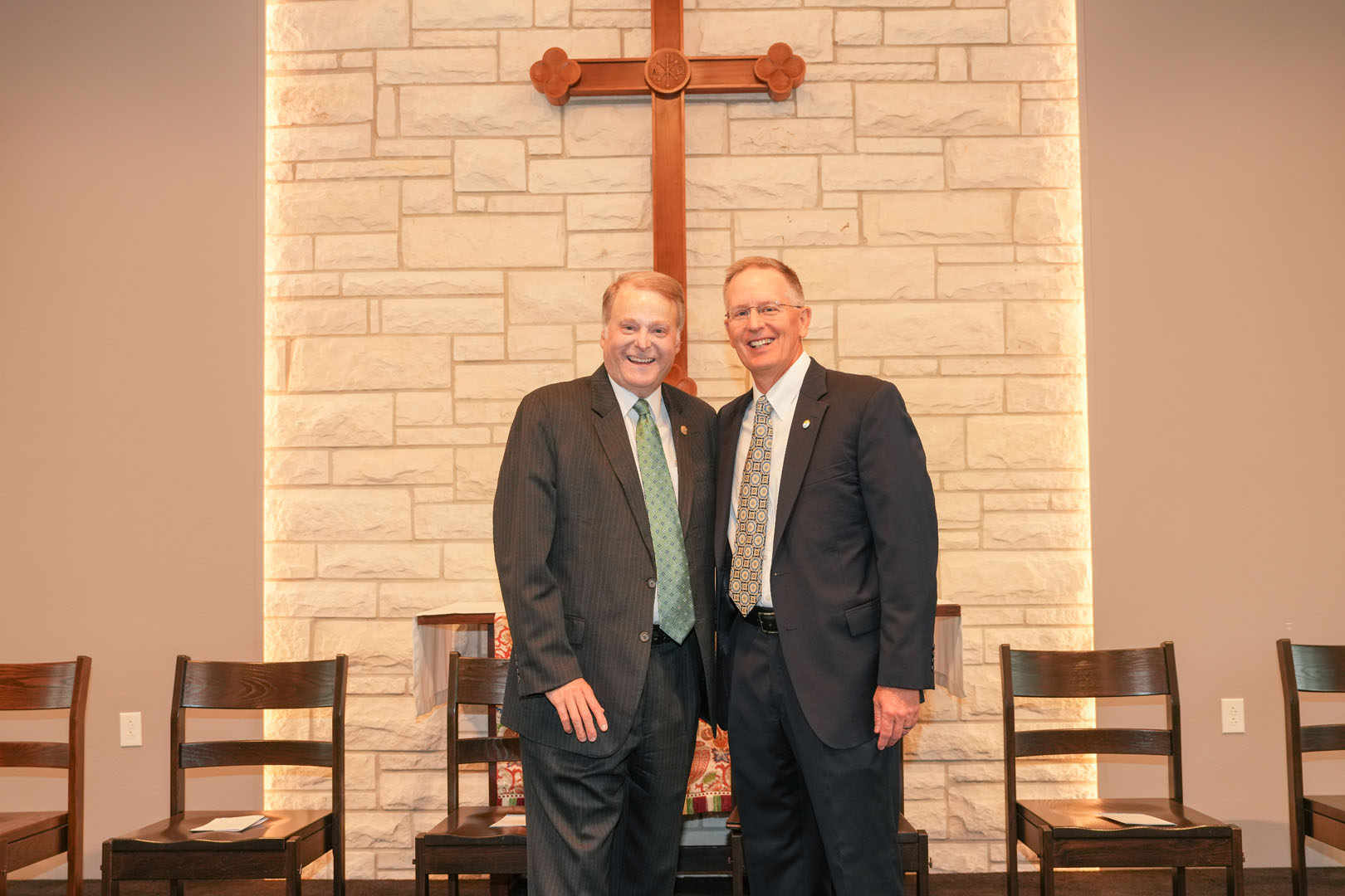 Two men in suits stand smiling in front of a wall with a large wooden cross mounted on it. The wall is made of light-colored bricks, and there are several chairs arranged in a row behind the men.