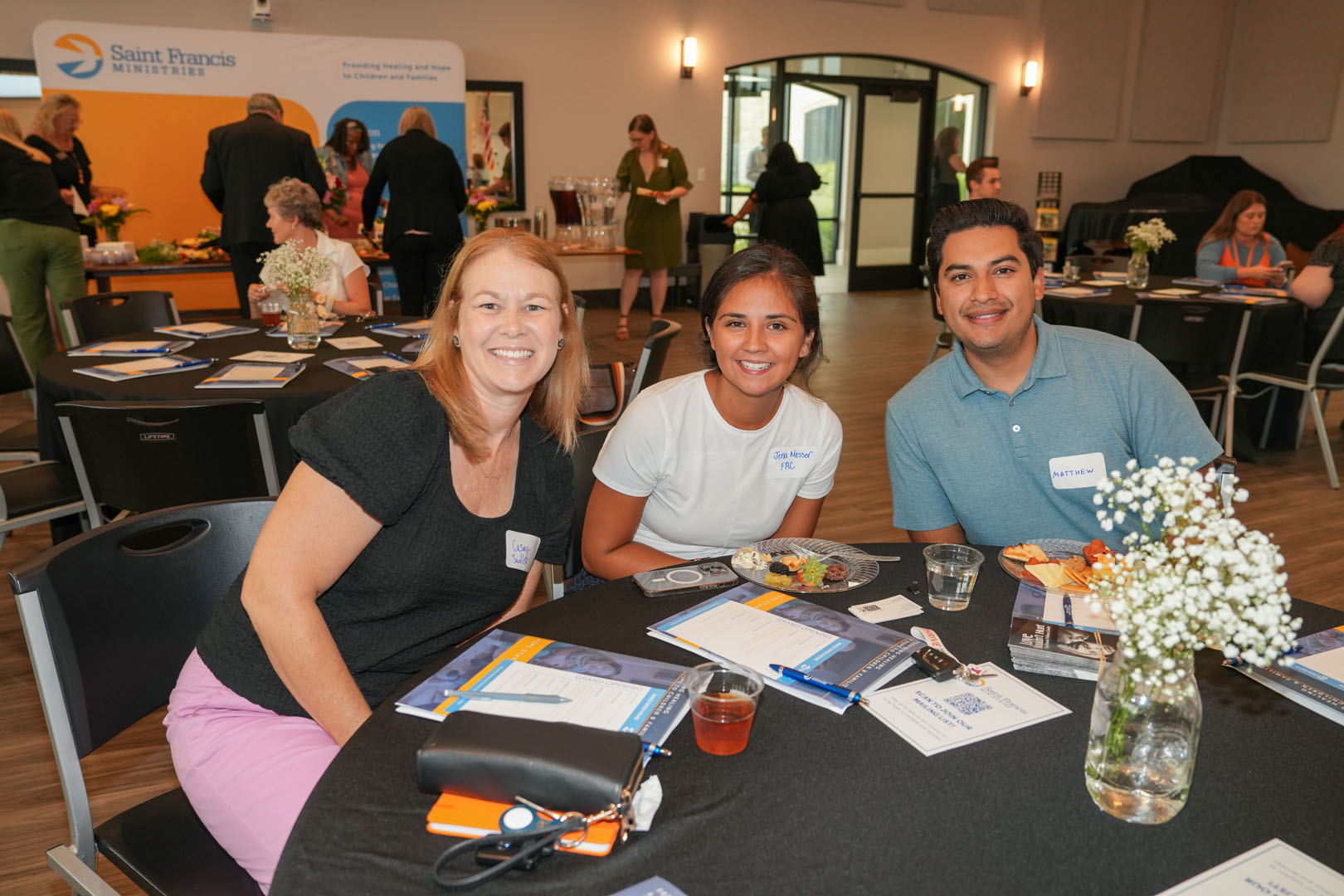 Three people are seated at a round table, smiling at the camera, in a room with Saint Francis Ministries banners. There are various papers, brochures, and a vase with flowers on the table. Other attendees are visible in the background, engaging in conversation.