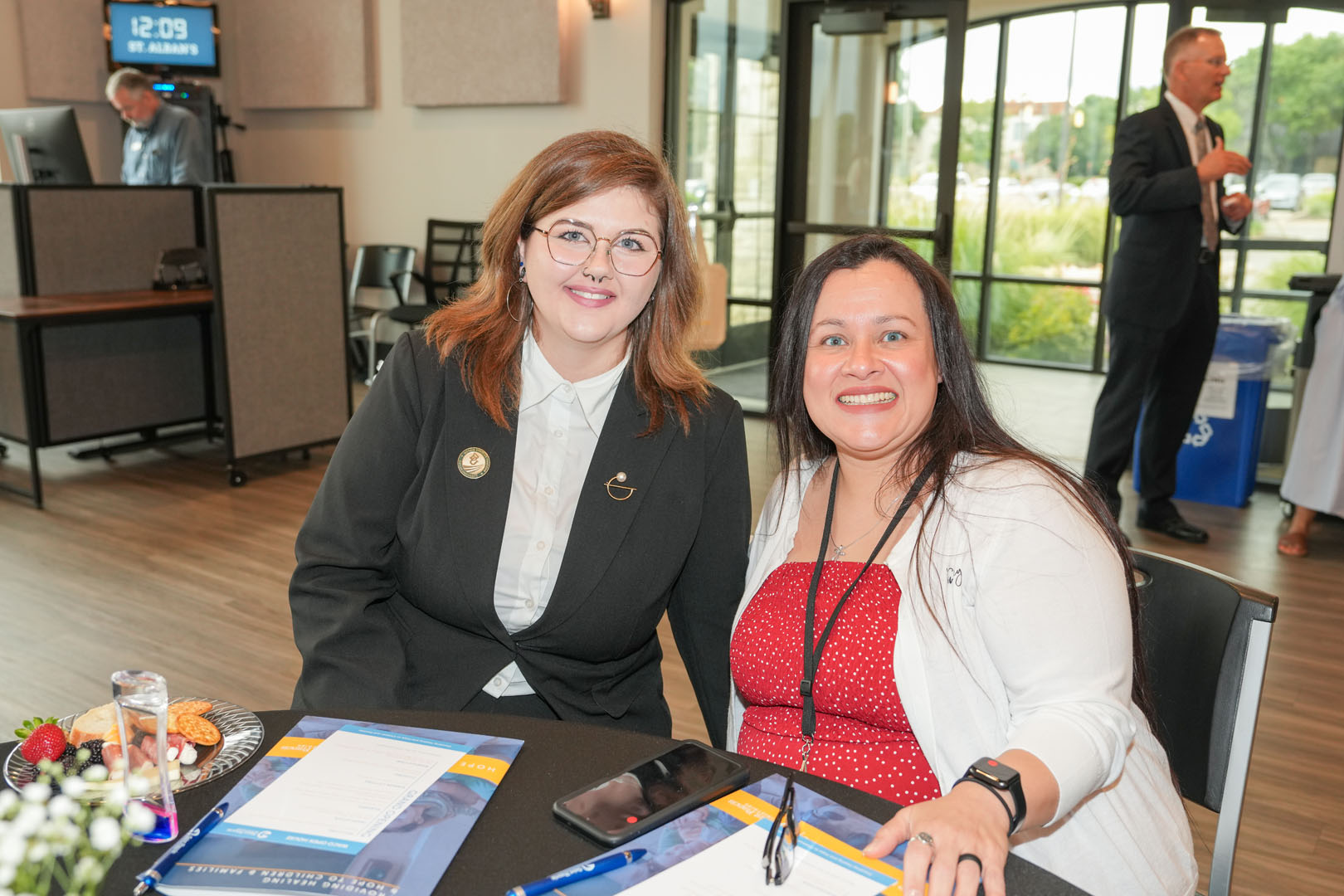 Two women are seated at a table inside a modern building. Both are smiling at the camera. The woman on the left is wearing a black blazer and glasses, while the woman on the right is wearing a white dress with a red cardigan. Papers and a phone are on the table.