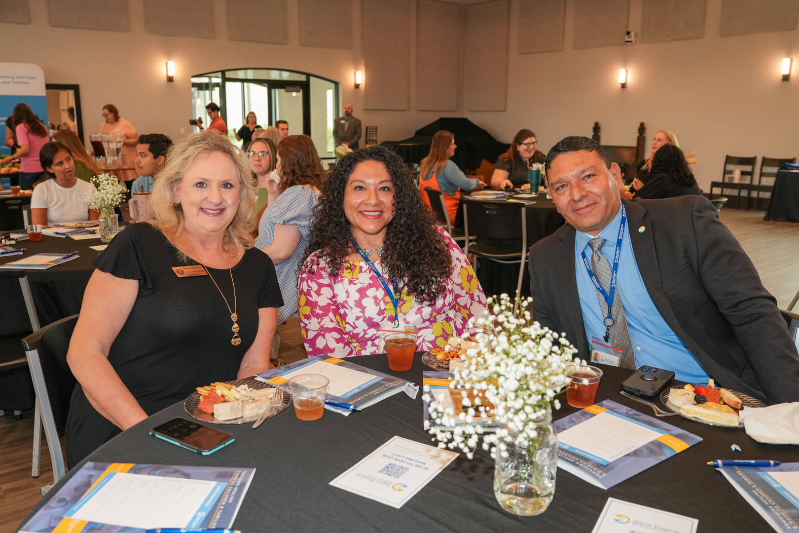 Three people sit at a round table during an event. They have plates with food and drinks in front of them and are smiling at the camera. Other attendees can be seen sitting at tables in the background. The atmosphere appears to be social and professional.