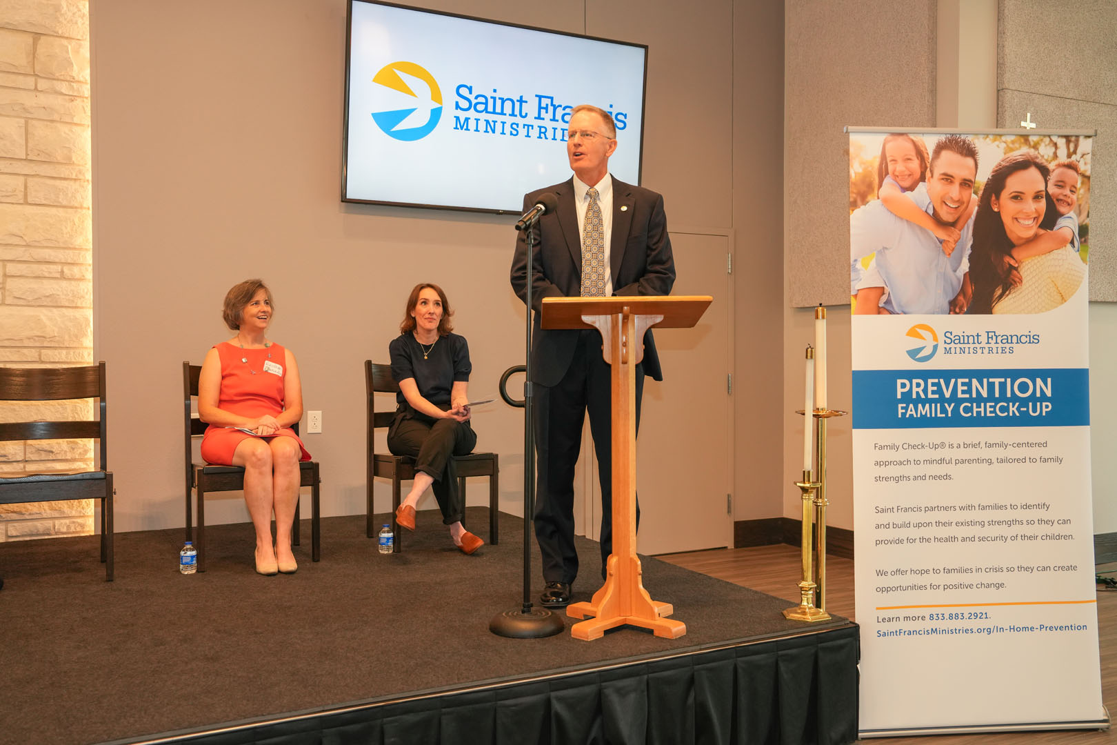 A man in a suit speaks at a podium with a "Saint Francis Ministries" banner nearby. Two women are seated on stage behind him, one wearing a red dress and the other in a blue suit. The scene appears to be a formal event or presentation.