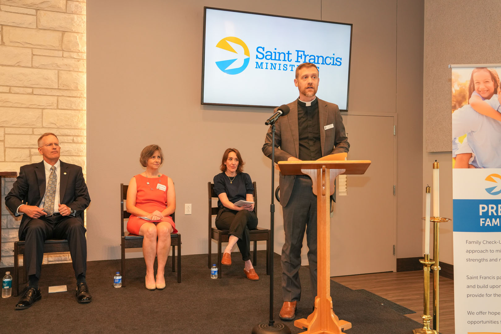 A man in a clerical collar stands at a podium speaking with a Saint Francis Ministries sign behind him. Three people are seated to his left, two men in business attire and a woman in a red dress. A vertical banner is on the right side of the image.