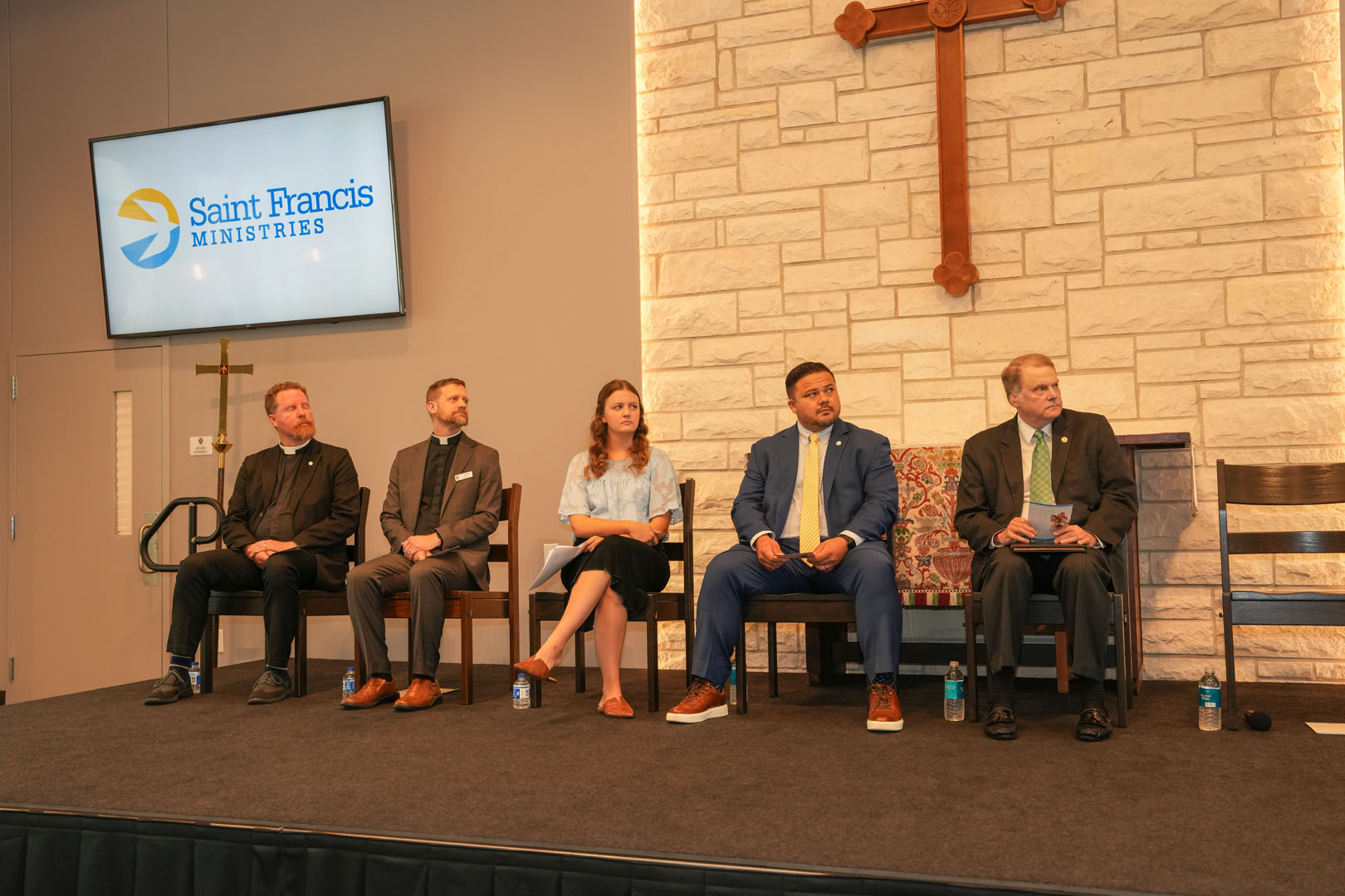 Five individuals are seated on a stage in front of a stone wall with a cross, under a screen displaying the logo "Saint Francis Ministries." They appear to be engaged in a panel discussion. Bottles of water are placed at their feet.