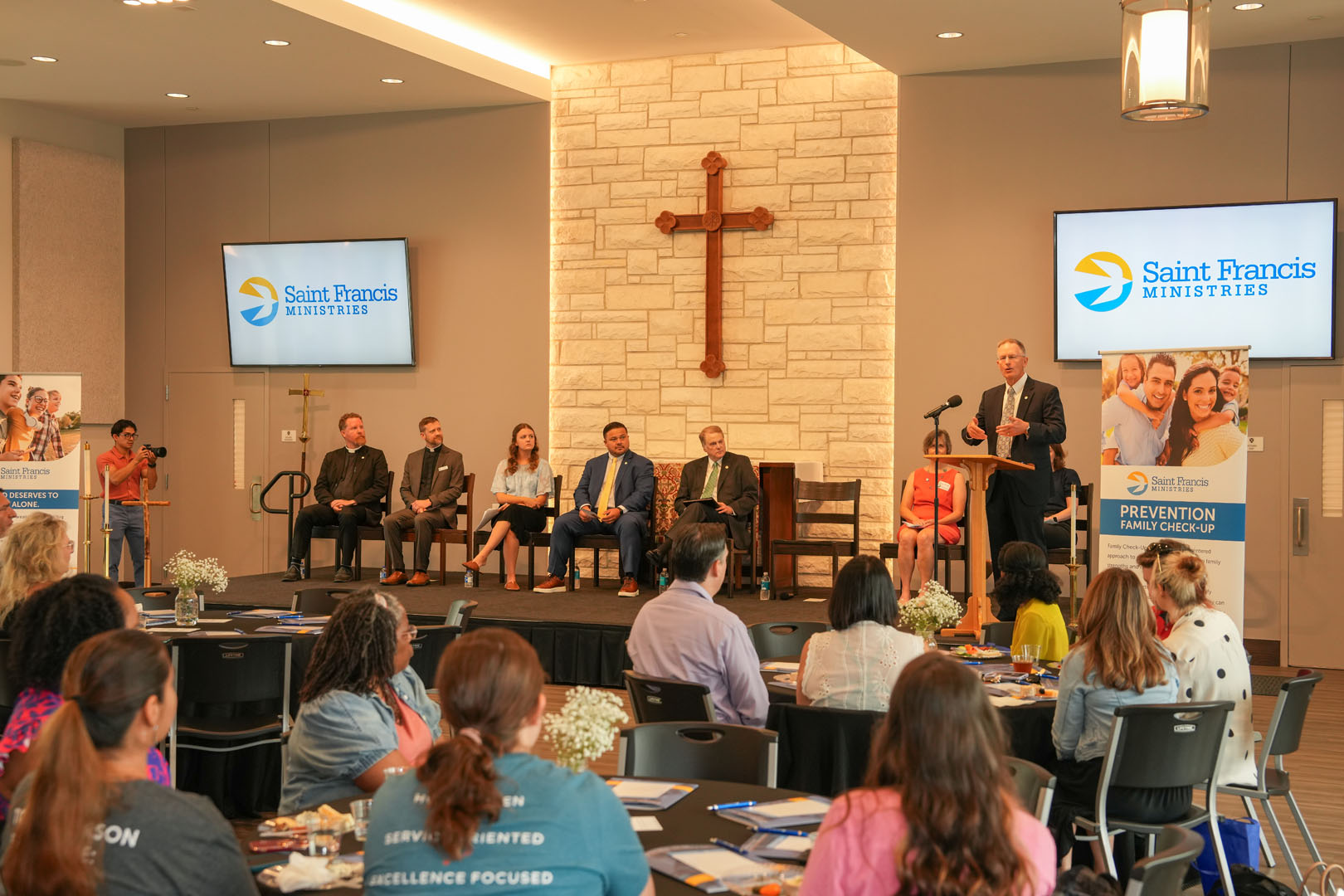 A panel of six people sits on a stage in front of a room of attendees at a Saint Francis Ministries event. There is a large wooden cross on the wall behind the panel. A speaker stands at a podium addressing the audience. Two projector screens display the event's logo.